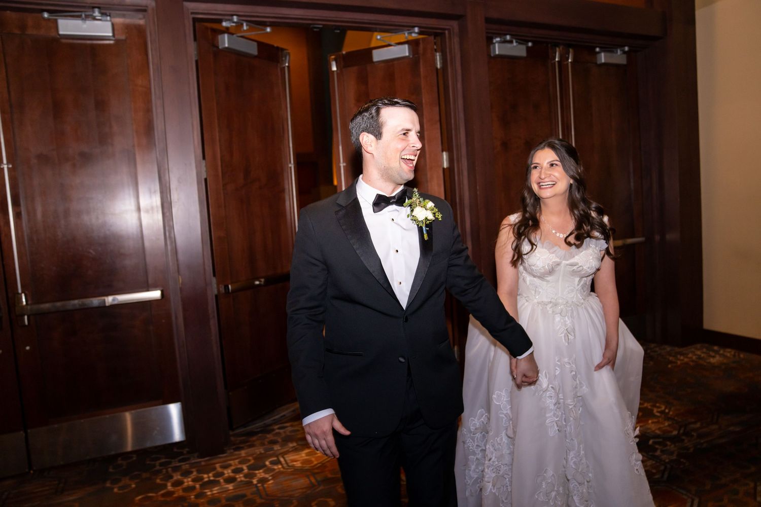 Smiling bride in white gown and groom in black tuxedo walking hand in hand through elegant hotel corridor.