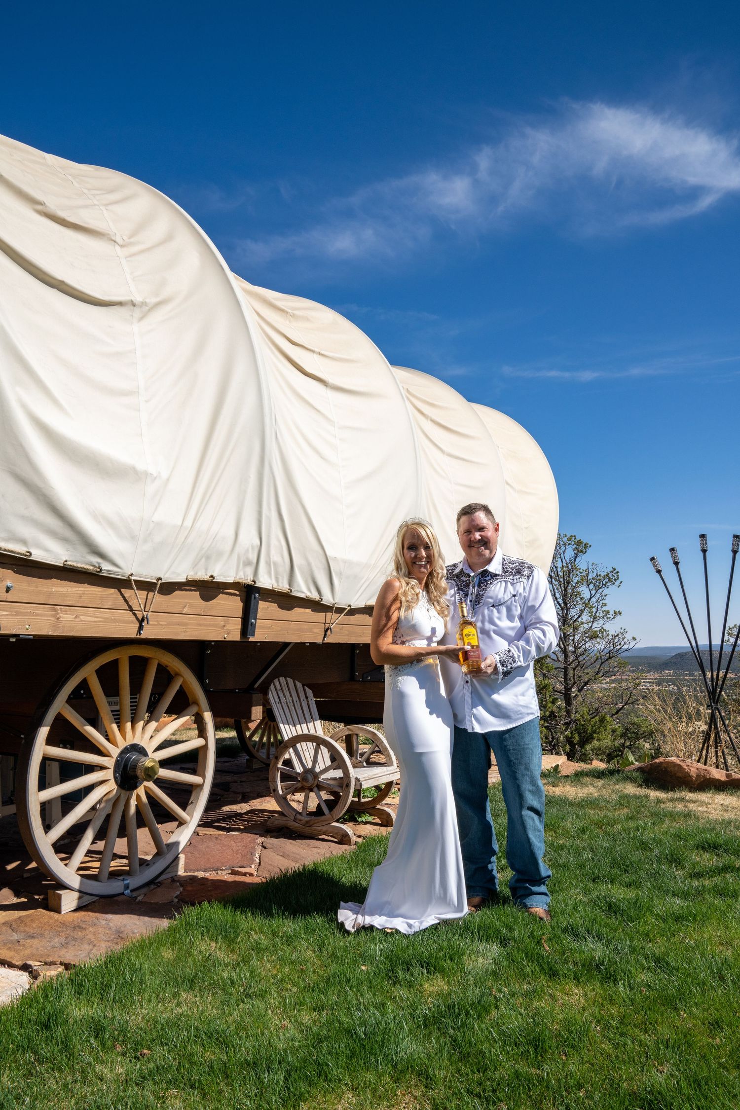 A couple poses for photos in front of a covered wagon at a rustic outdoor venue against a bright blue sky.