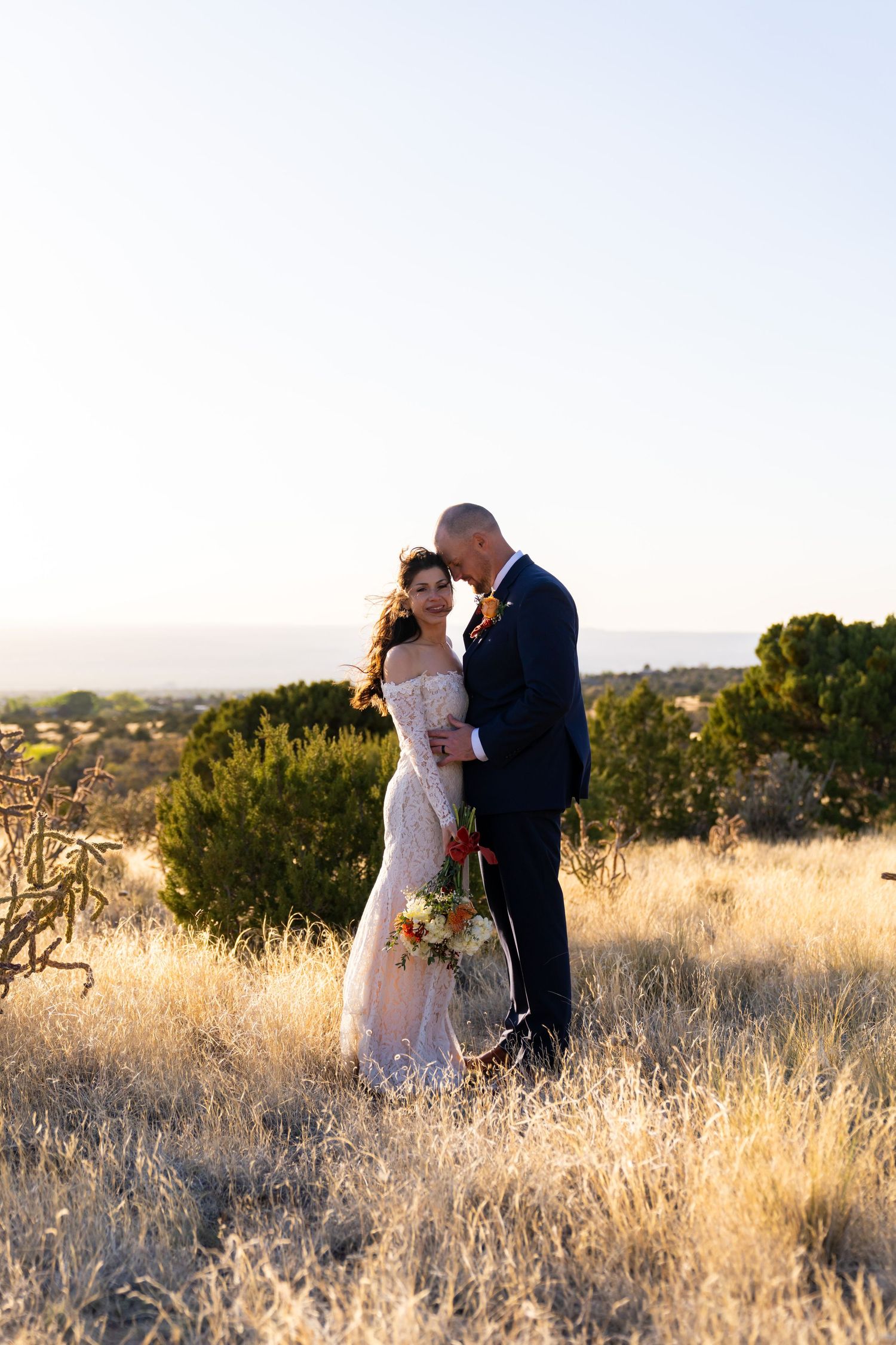 Bride and groom embrace in golden dry grass field at sunset during outdoor desert wedding portrait.