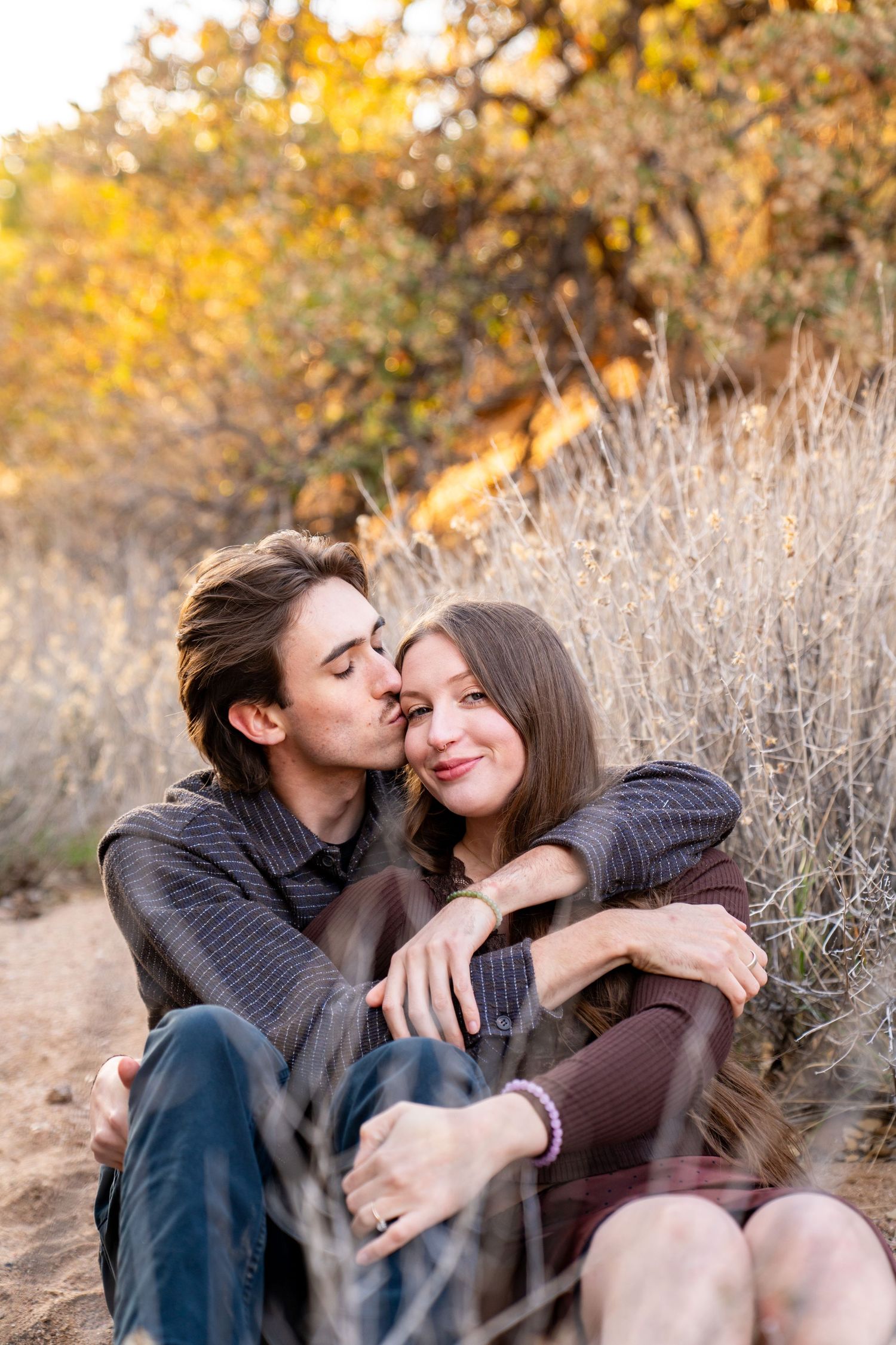 A couple sits together outdoors in autumn, sharing a tender kiss on the cheek amid golden dry grass and fall foliage.