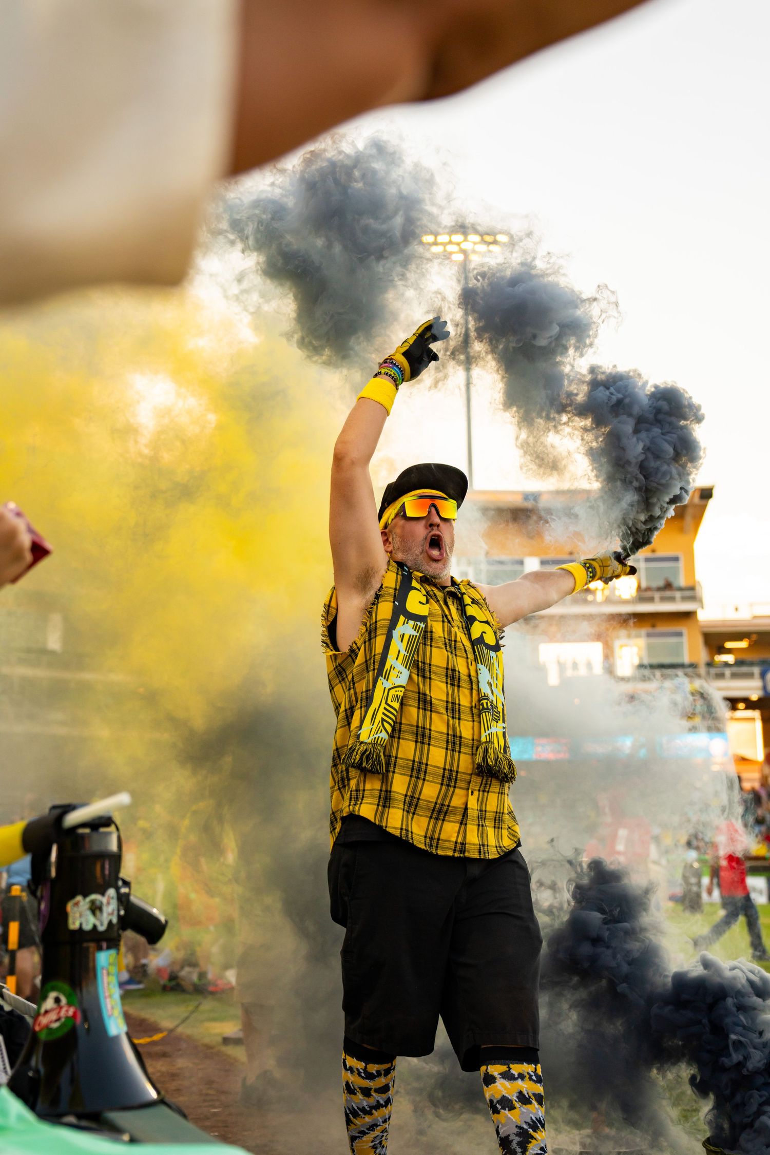Energetic soccer fan in yellow plaid shirt waves black and yellow smoke flares at a stadium celebration.