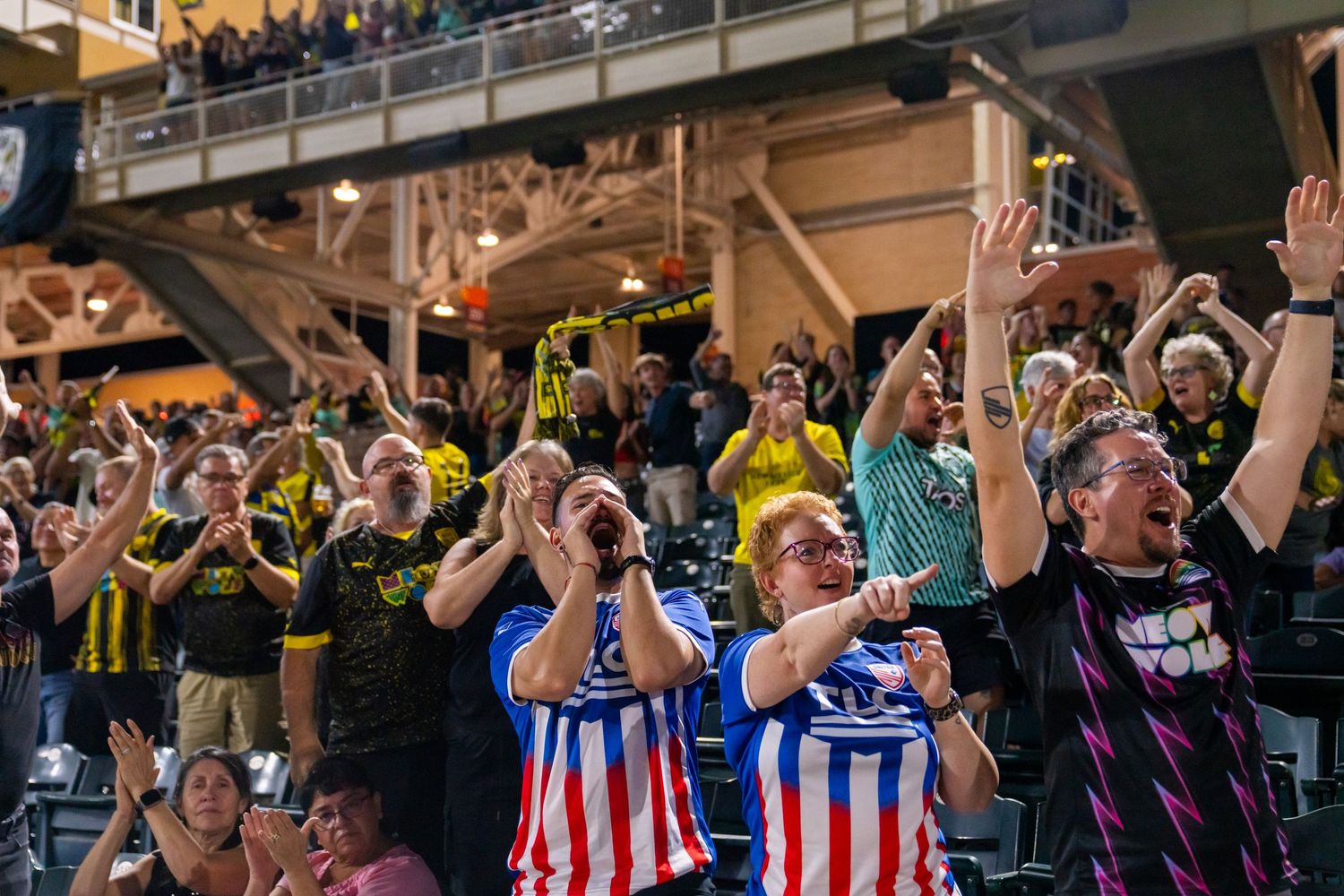 Excited soccer fans cheering in stadium stands, wearing team jerseys in black and blue, celebrating during a live match.