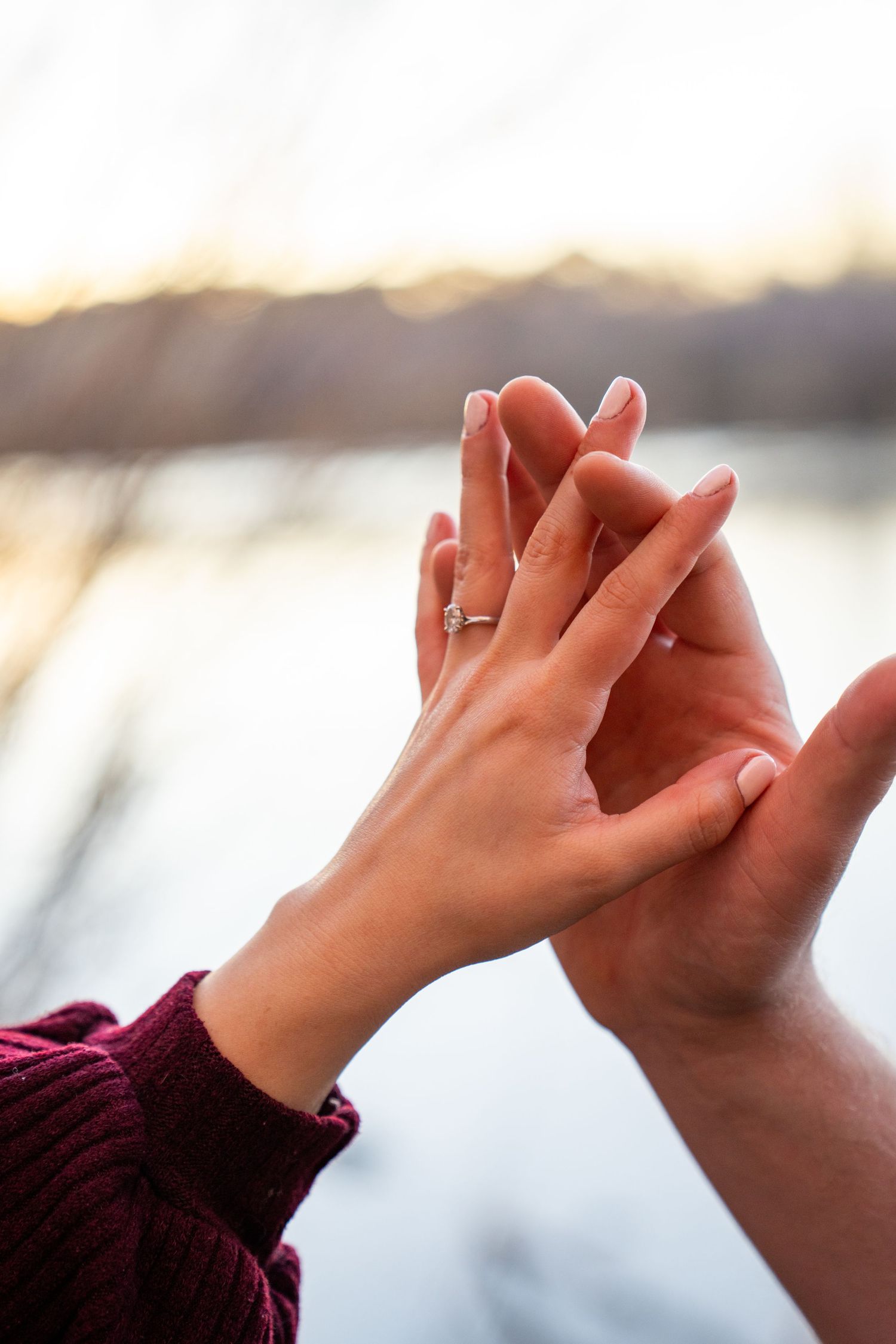 Close-up of two hands intertwined, woman wearing a diamond engagement ring, romantic sunset by a lake.