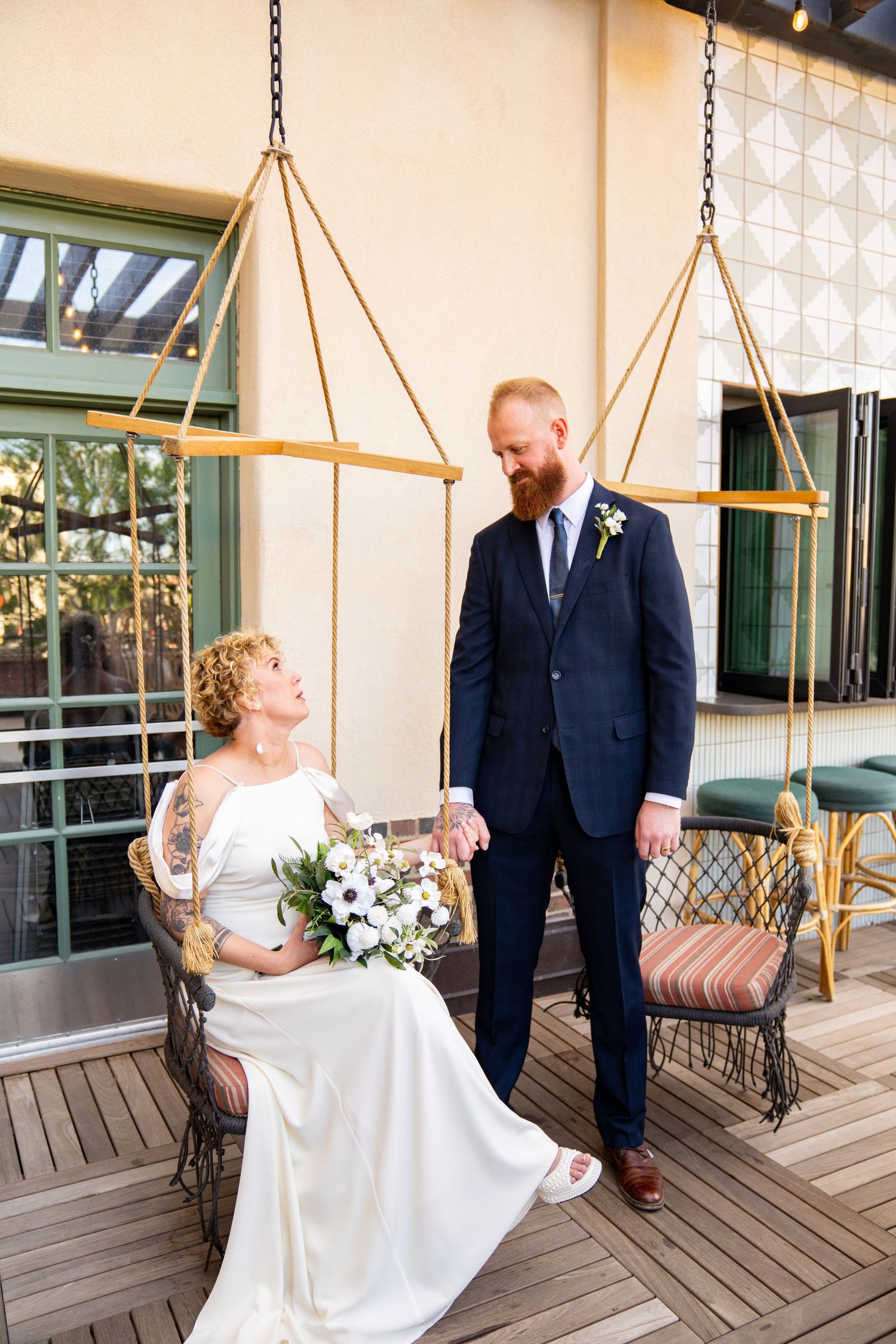A bride in a white gown holds a bouquet while seated as her groom in a navy suit stands beside her on a wooden deck.