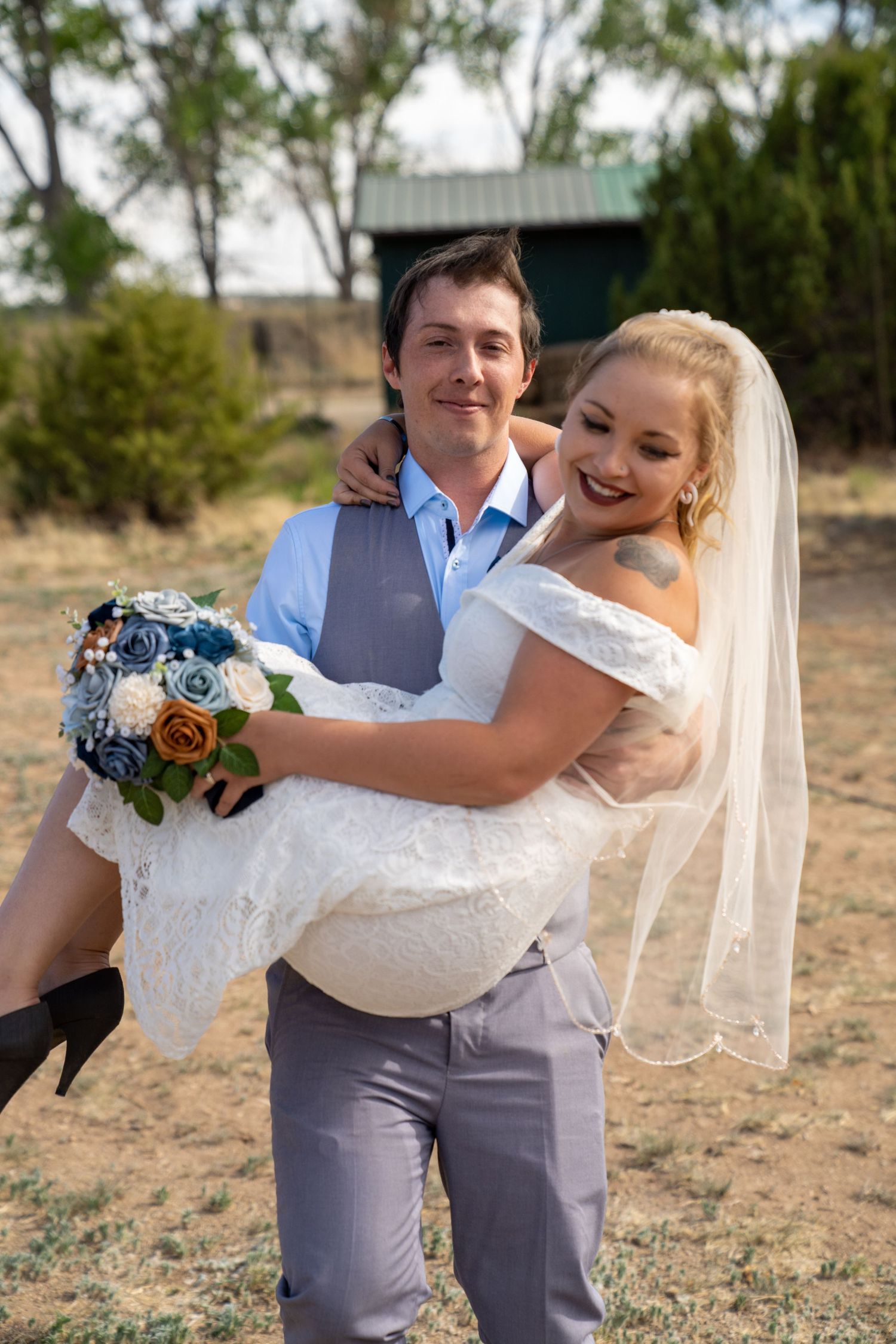 A couple shares a playful moment outdoors with the groom carrying the bride in her white lace dress.