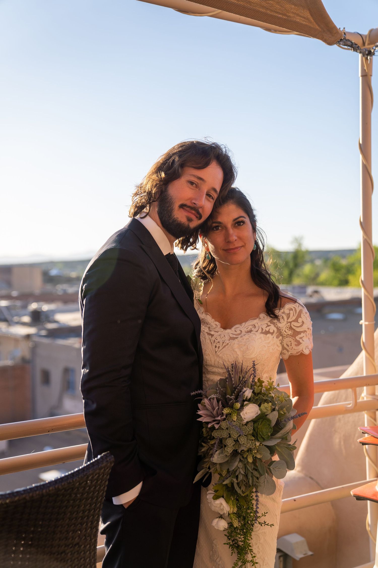 A couple sharing a tender moment on a rooftop terrace under a wooden pergola against a bright blue sky.