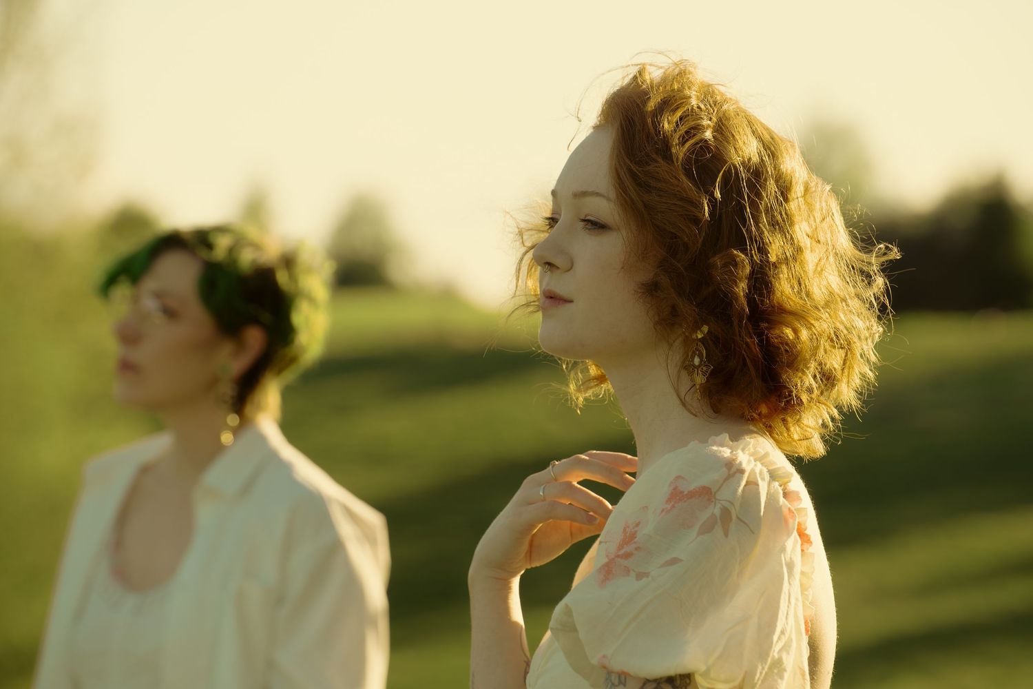 Two women in white dresses outdoors, one with a flower crown, bathed in warm golden sunlight.