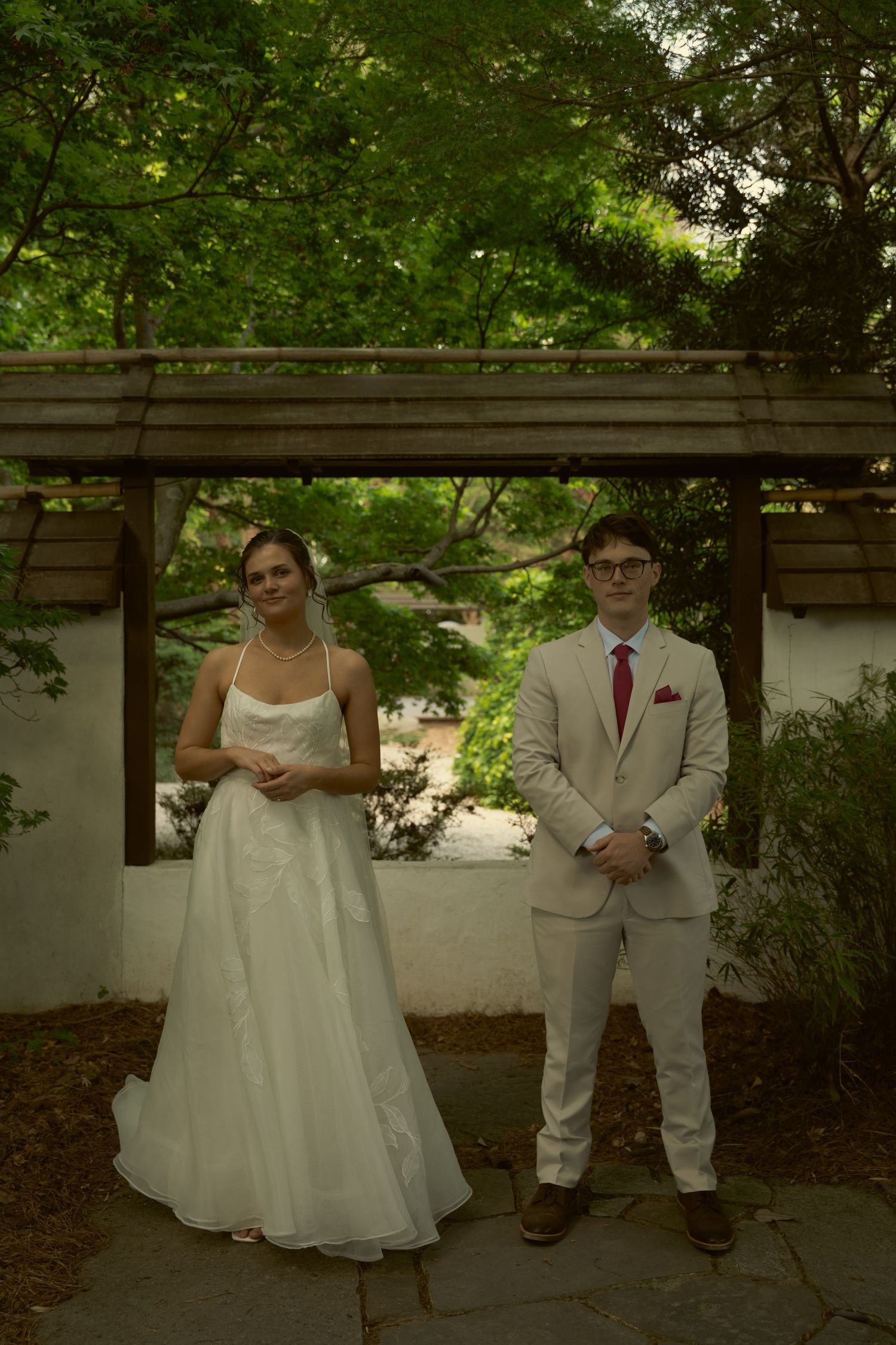 Bride and groom posing together under a wooden pergola surrounded by lush green trees.
