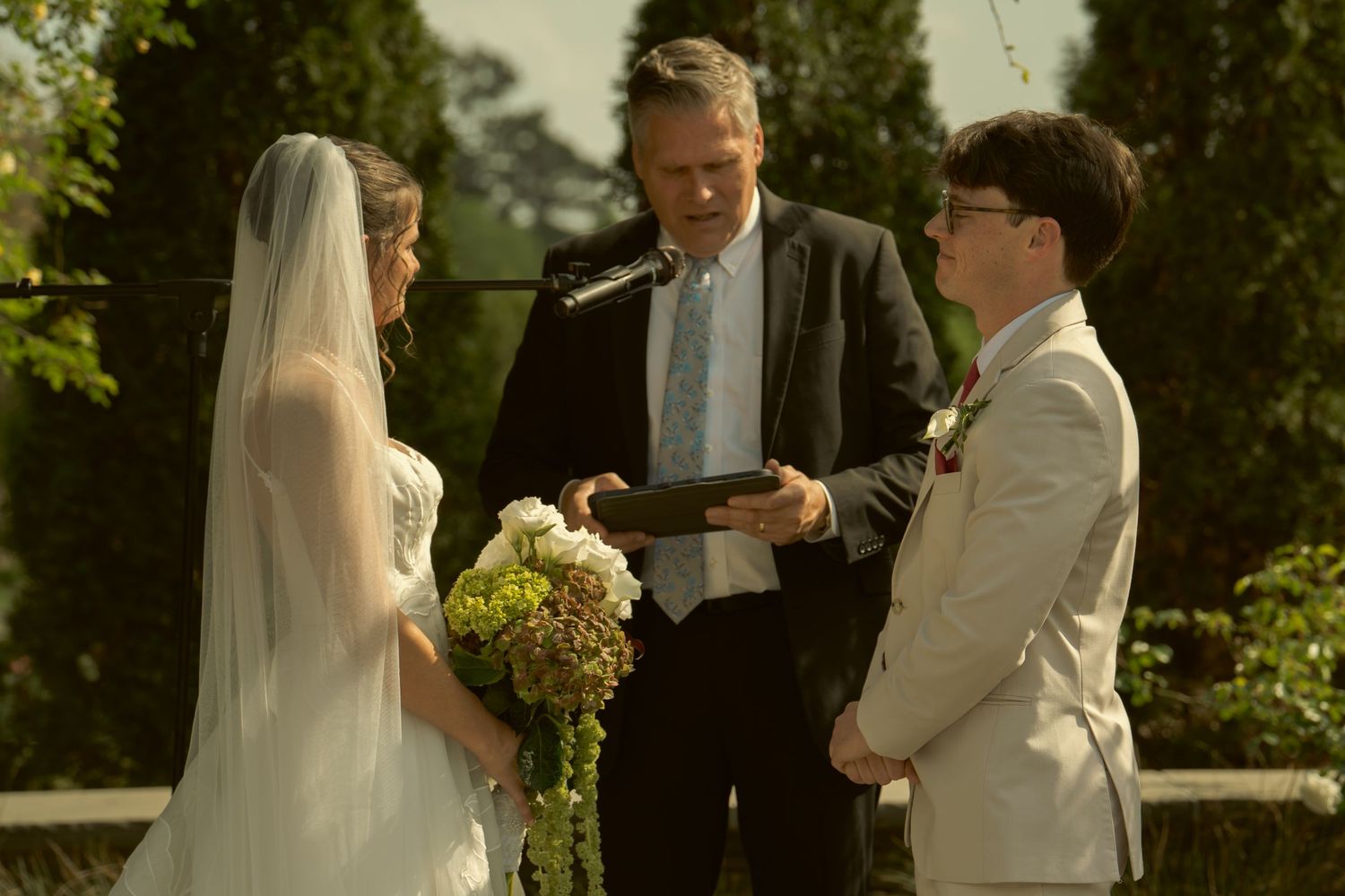 Wedding ceremony outdoors, officiant reading vows as bride in white veil and groom in tan suit exchange rings.