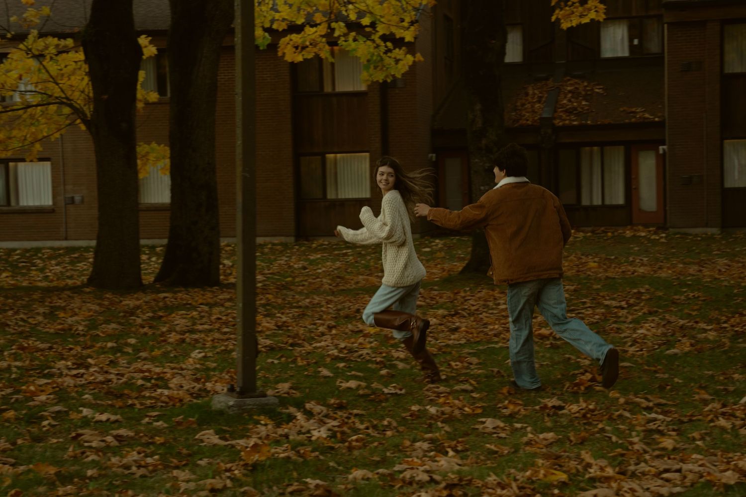 Figures walking among trees in a park with fallen autumn leaves.