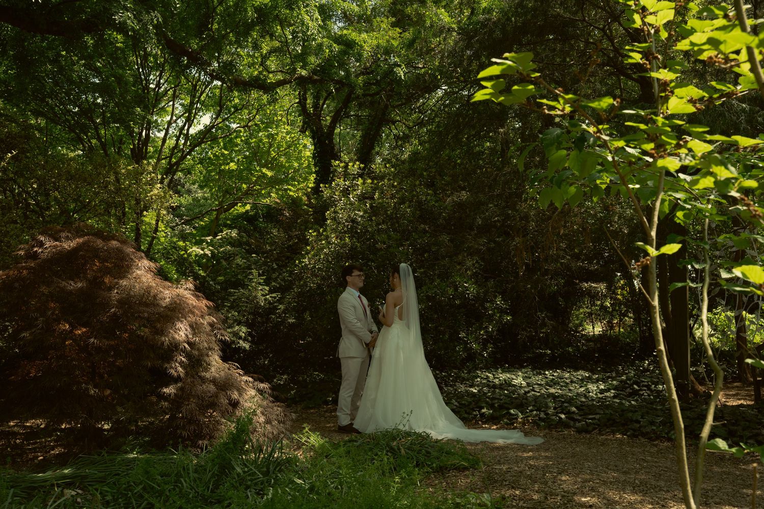 Bride and groom stand hand in hand in sunlit forest garden during outdoor wedding ceremony.