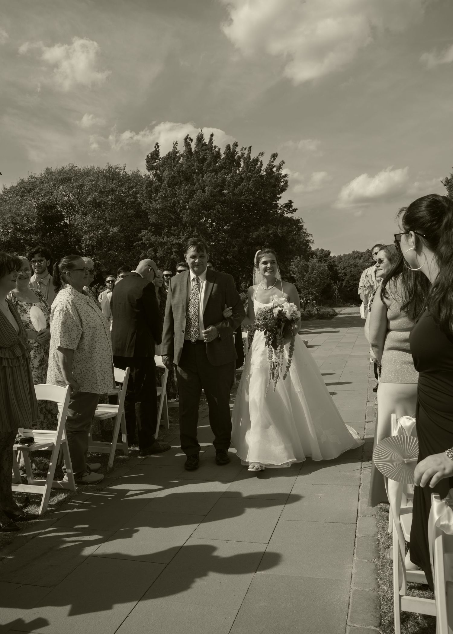 Black and white photo of a bride walking down the outdoor aisle with a man at a sunny wedding ceremony.