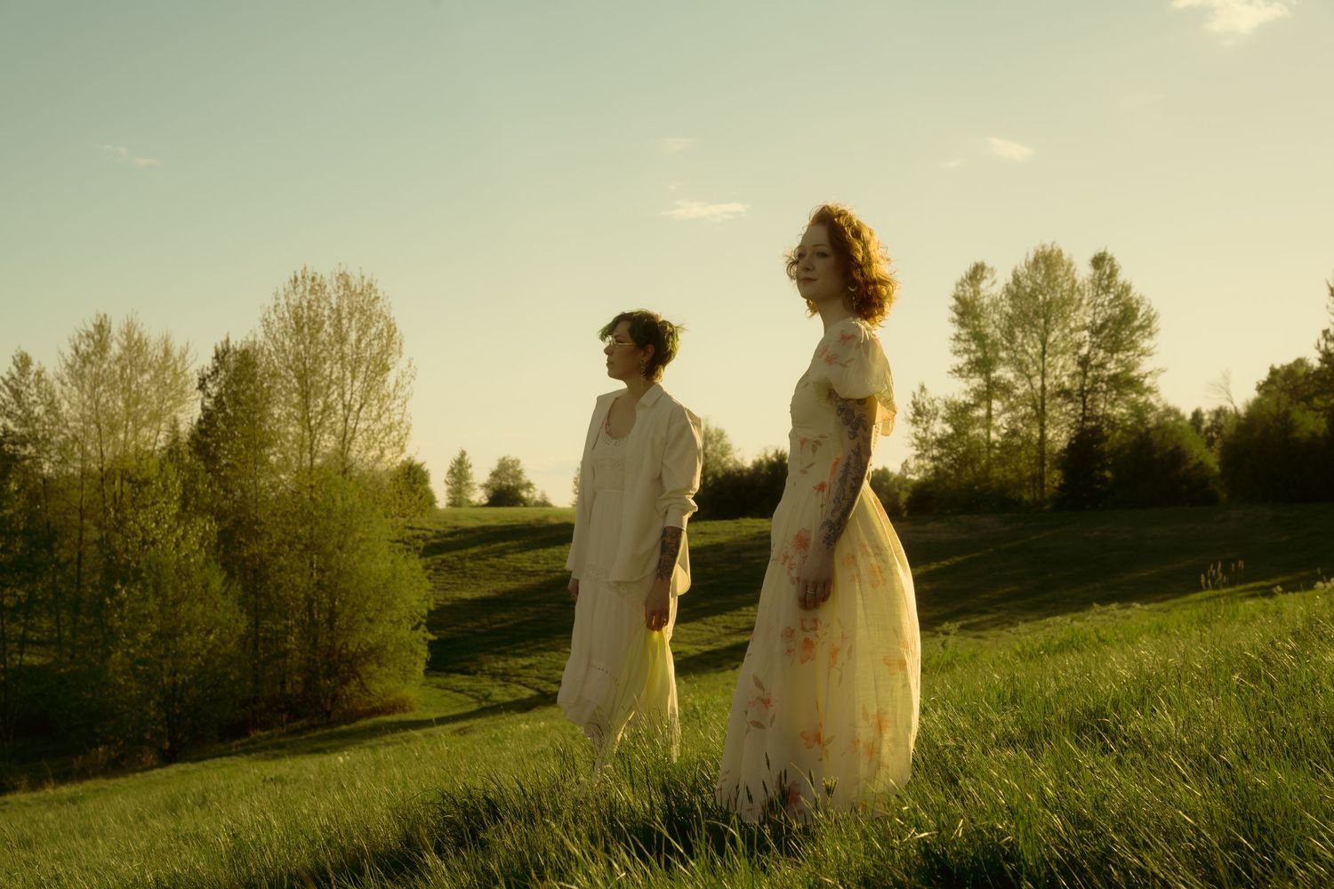 Couple in vintage white garments stand together on lush green hillside during warm sunset light.