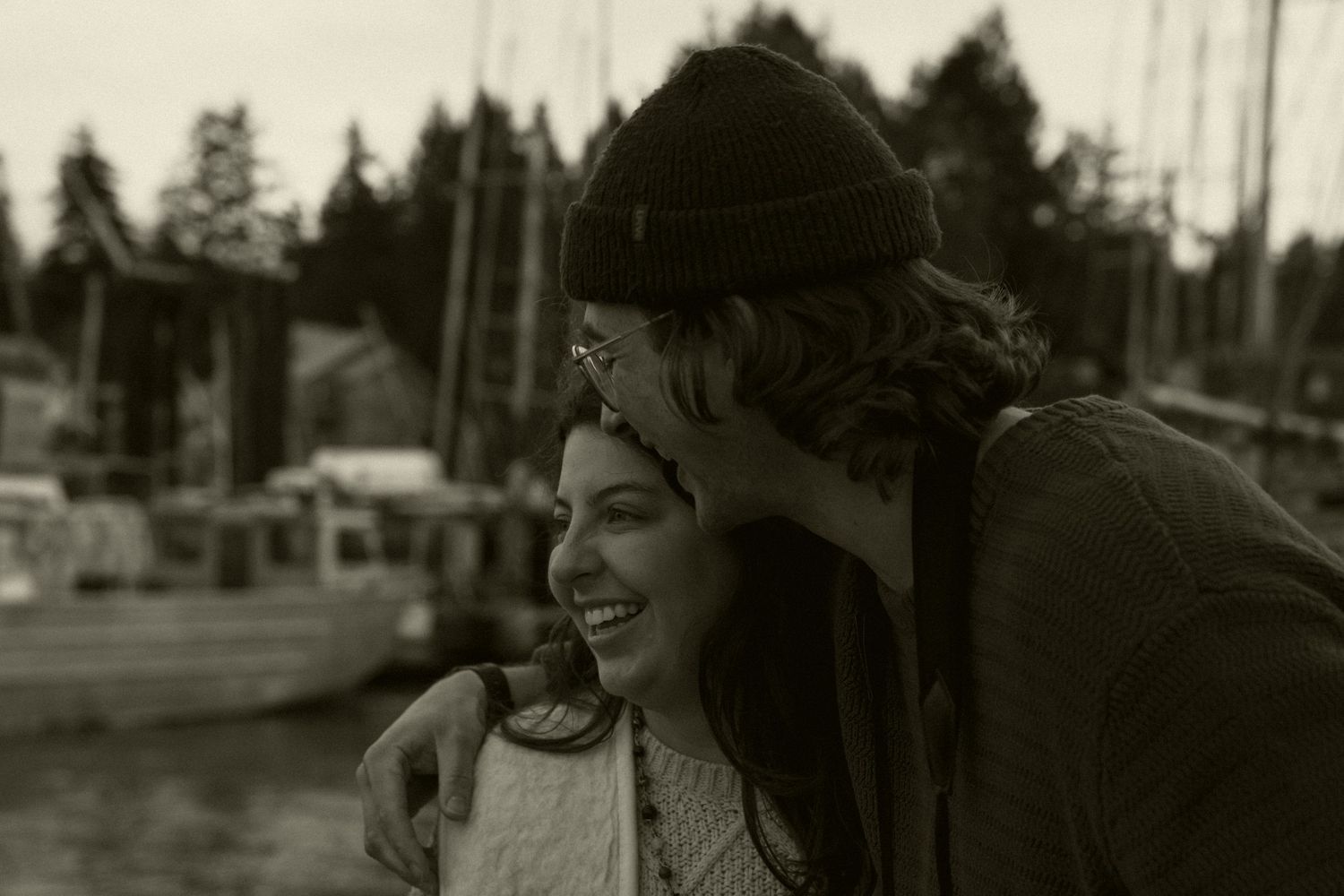 Couple sharing a tender moment at a marina, laughing together in black and white photo with boats in background.