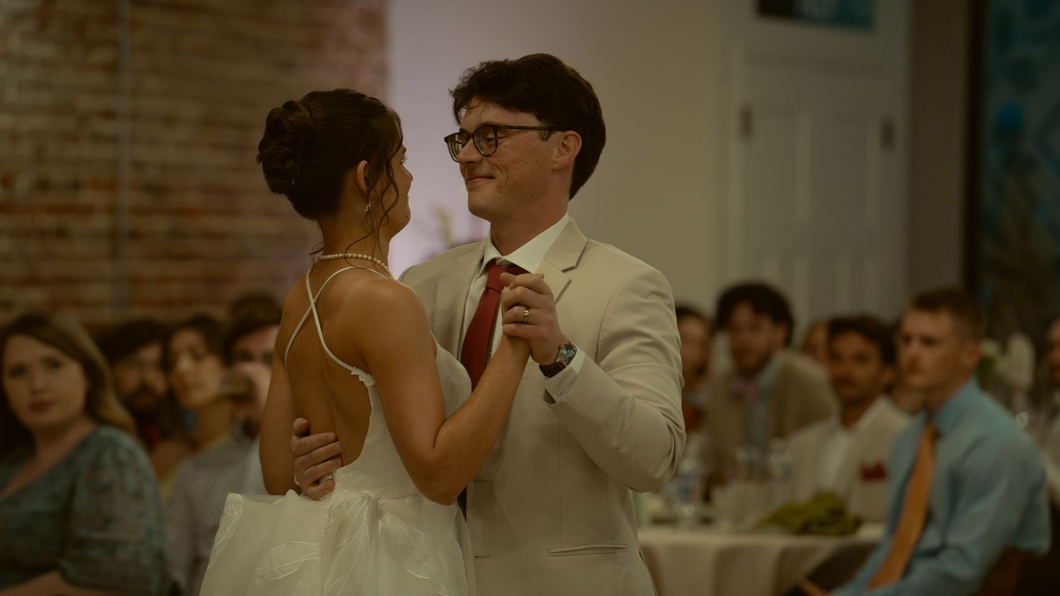 Couple sharing first dance at wedding reception in brick venue, groom in white suit with red tie.