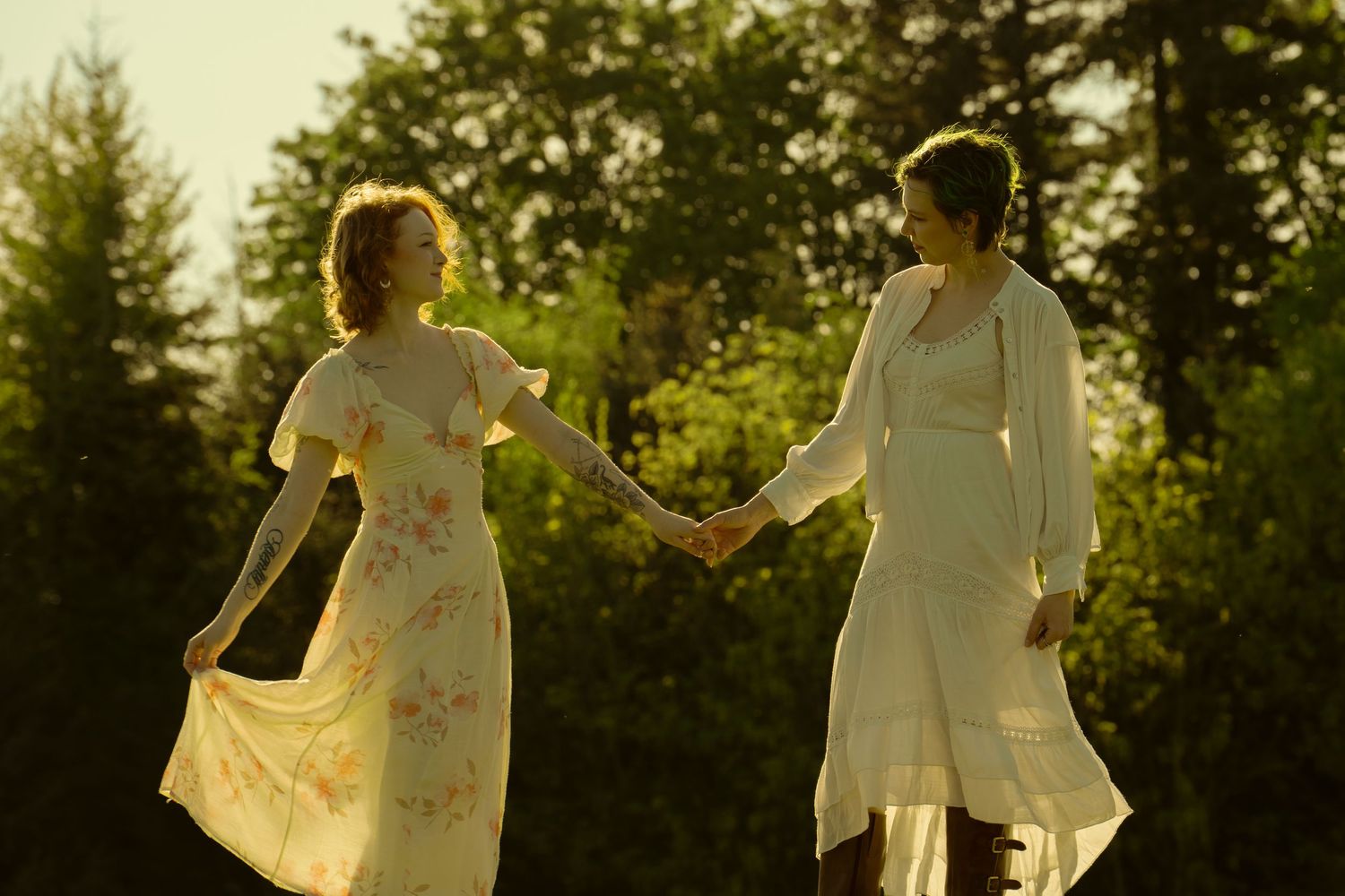 Two women in flowing floral dresses hold hands outdoors in golden sunlight with green trees behind them.