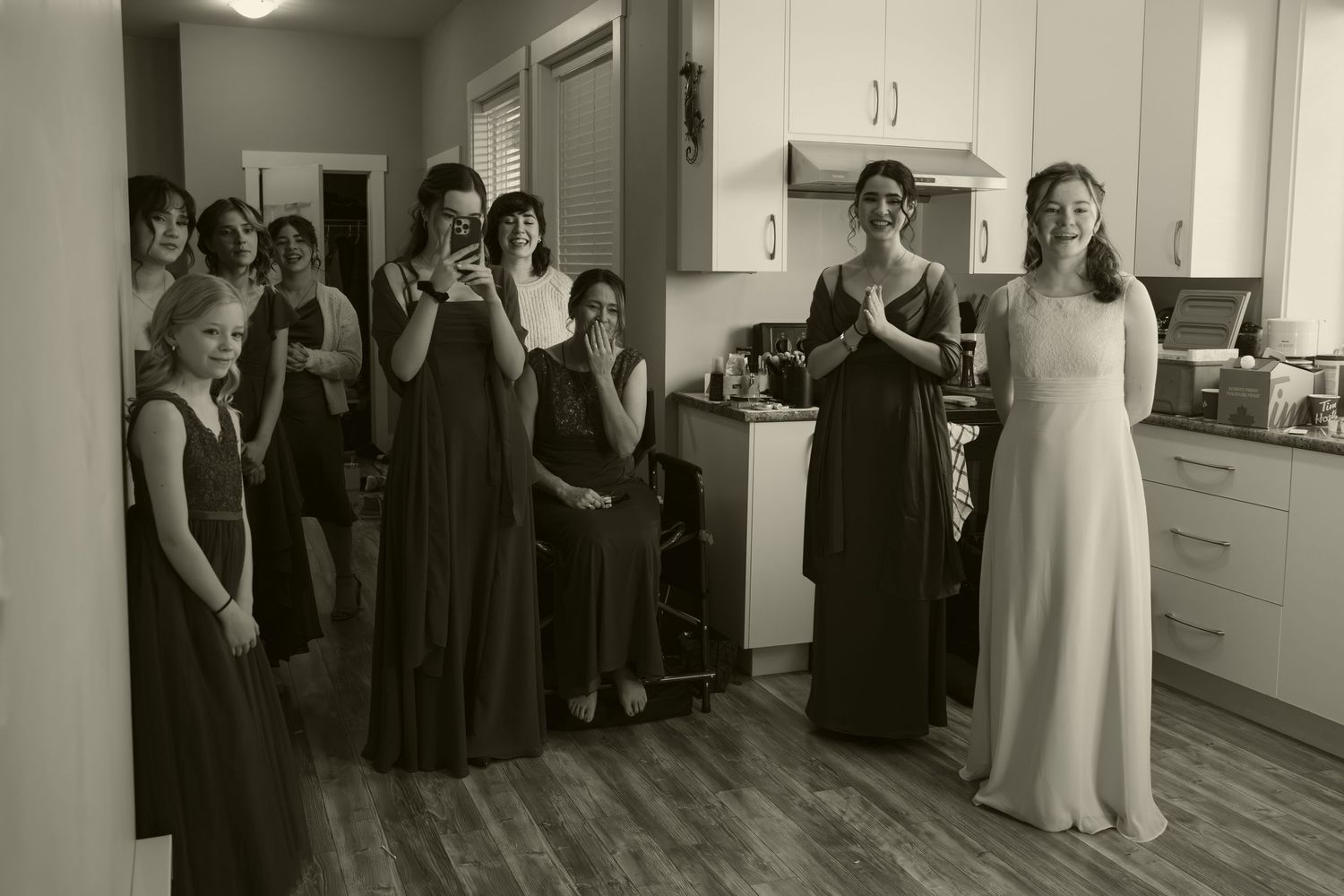 A group wearing formal black dresses and a white gown gather in a kitchen during wedding preparations.