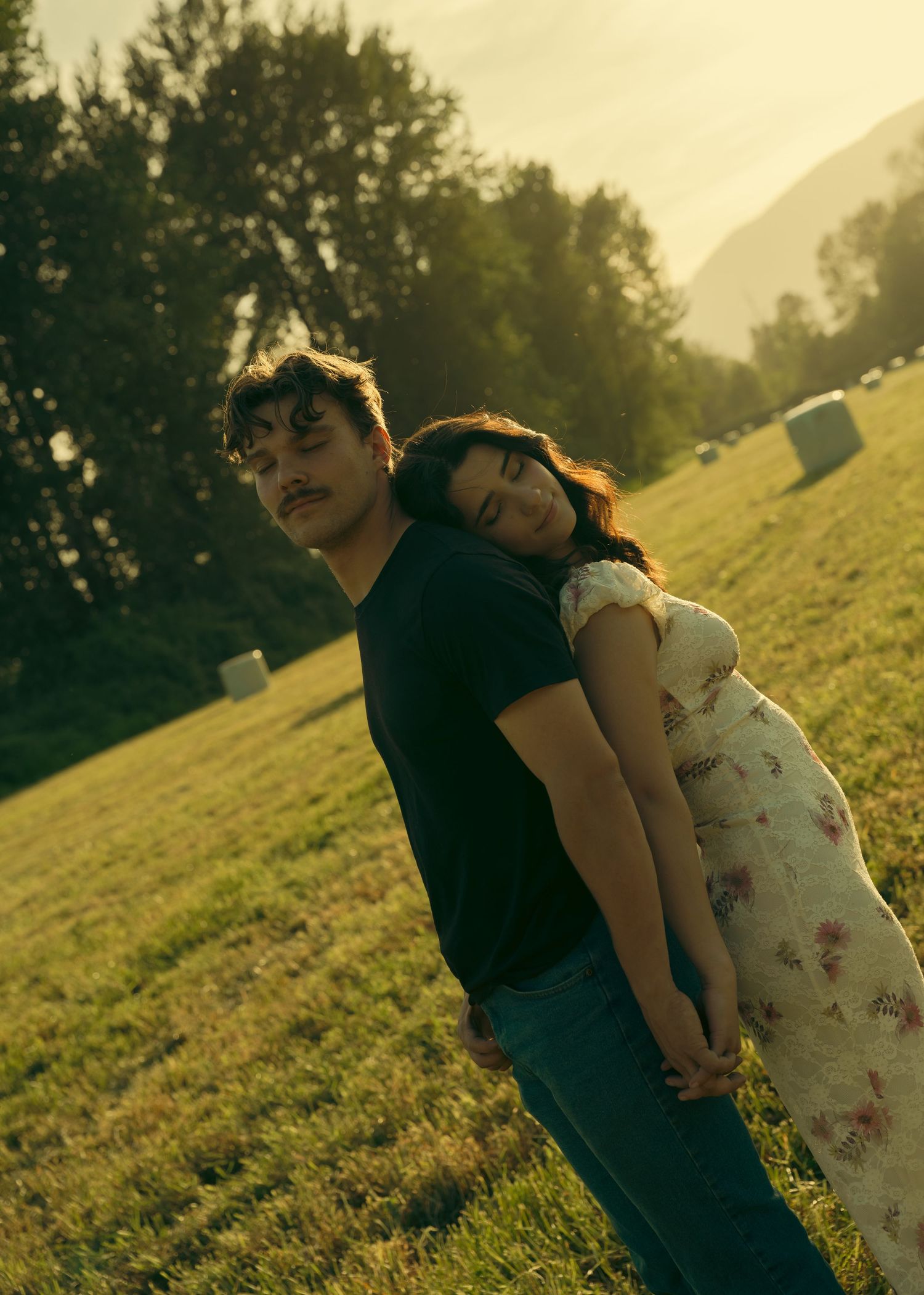 A young couple shares a tender moment in a golden sunlit field, the woman leaning her head on the man's back.