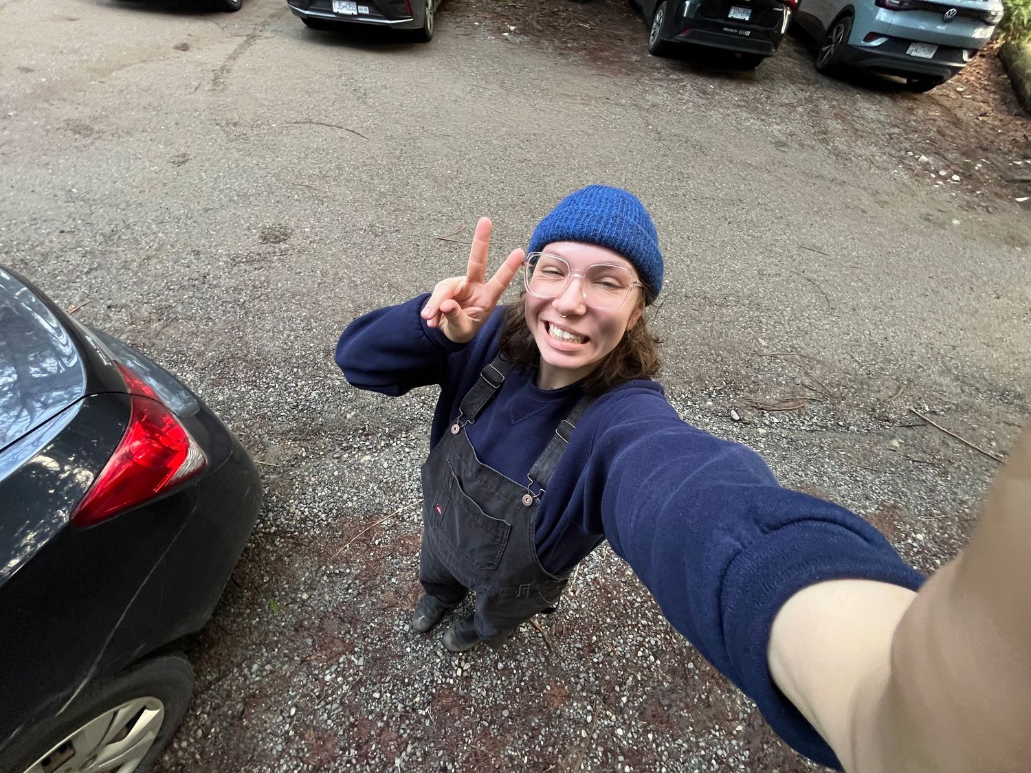 A selfie taken in a parking lot showing someone wearing a blue beanie and navy jacket making a peace sign.