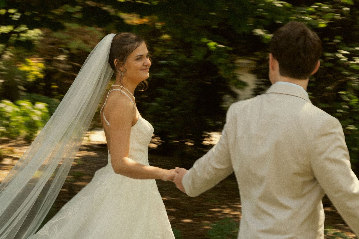 Smiling bride in veil holds grooms hand while walking through a sunlit garden.