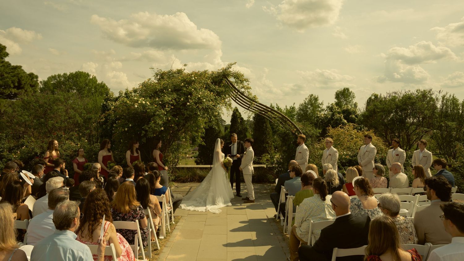 Outdoor wedding ceremony with guests seated under a floral arch, surrounded by lush greenery on a partly cloudy day.