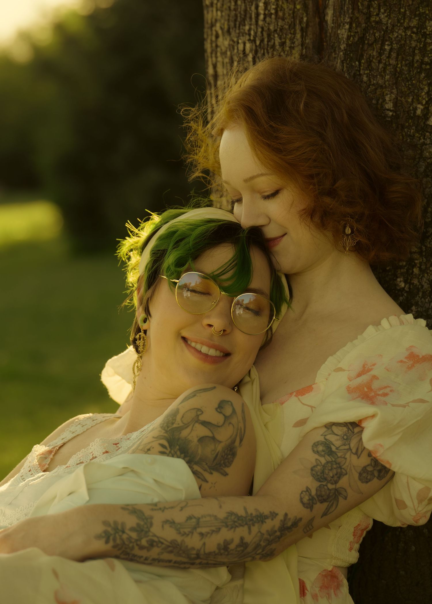 Two women embrace tenderly by a tree, one with green hair and tattoos, bathed in warm sunlight.