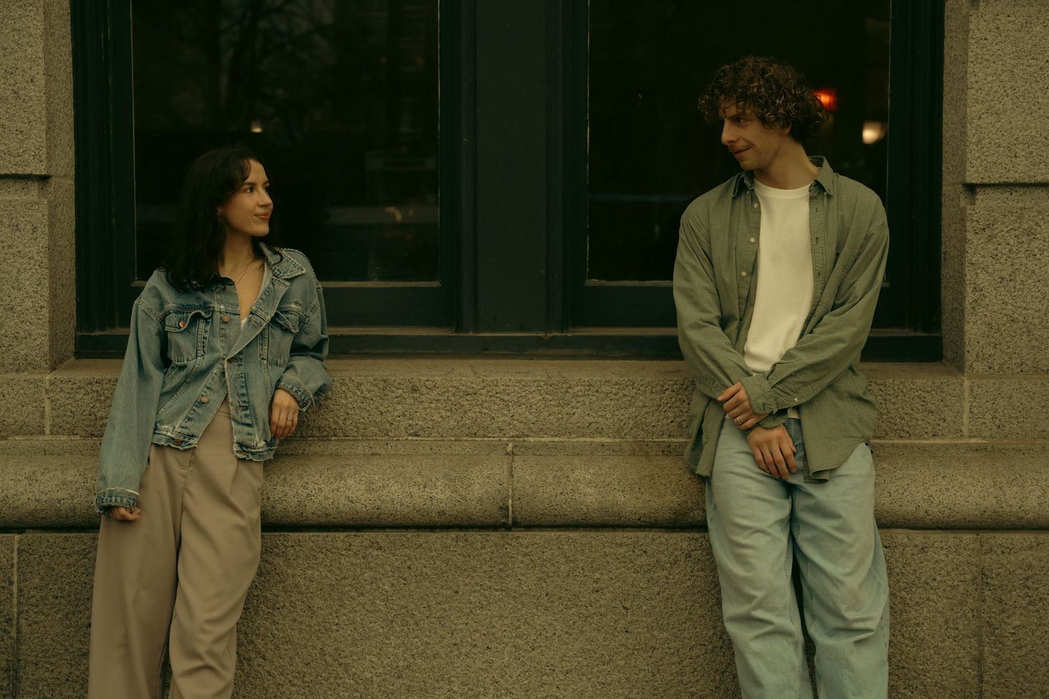 Two people in casual 90s style clothing stand near a building entrance.