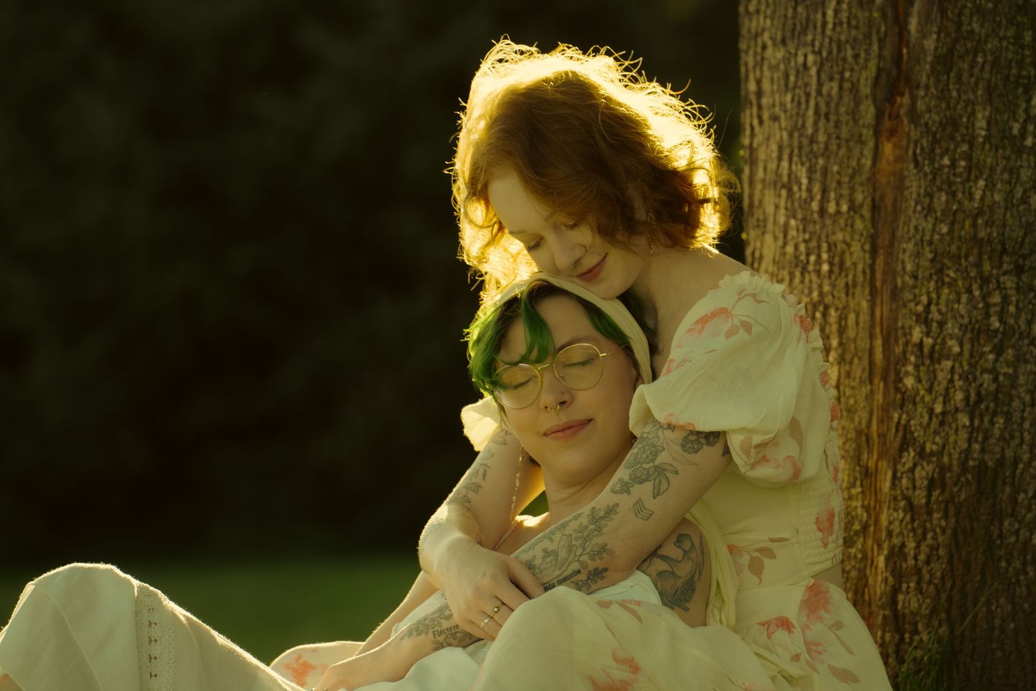 Two women embracing tenderly by a tree in warm golden light.