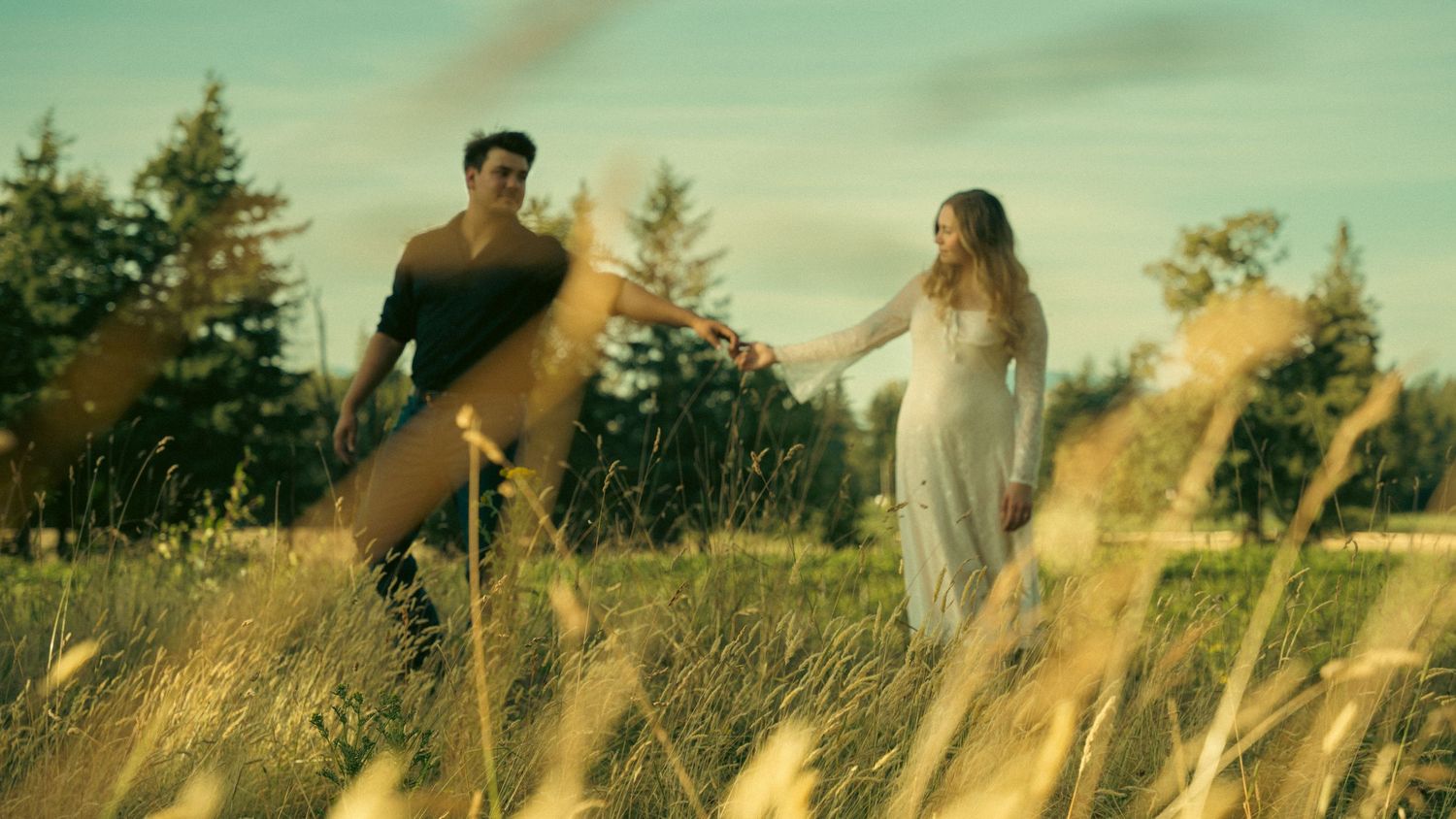 Couple walking through a sunlit meadow with mountains in the background.