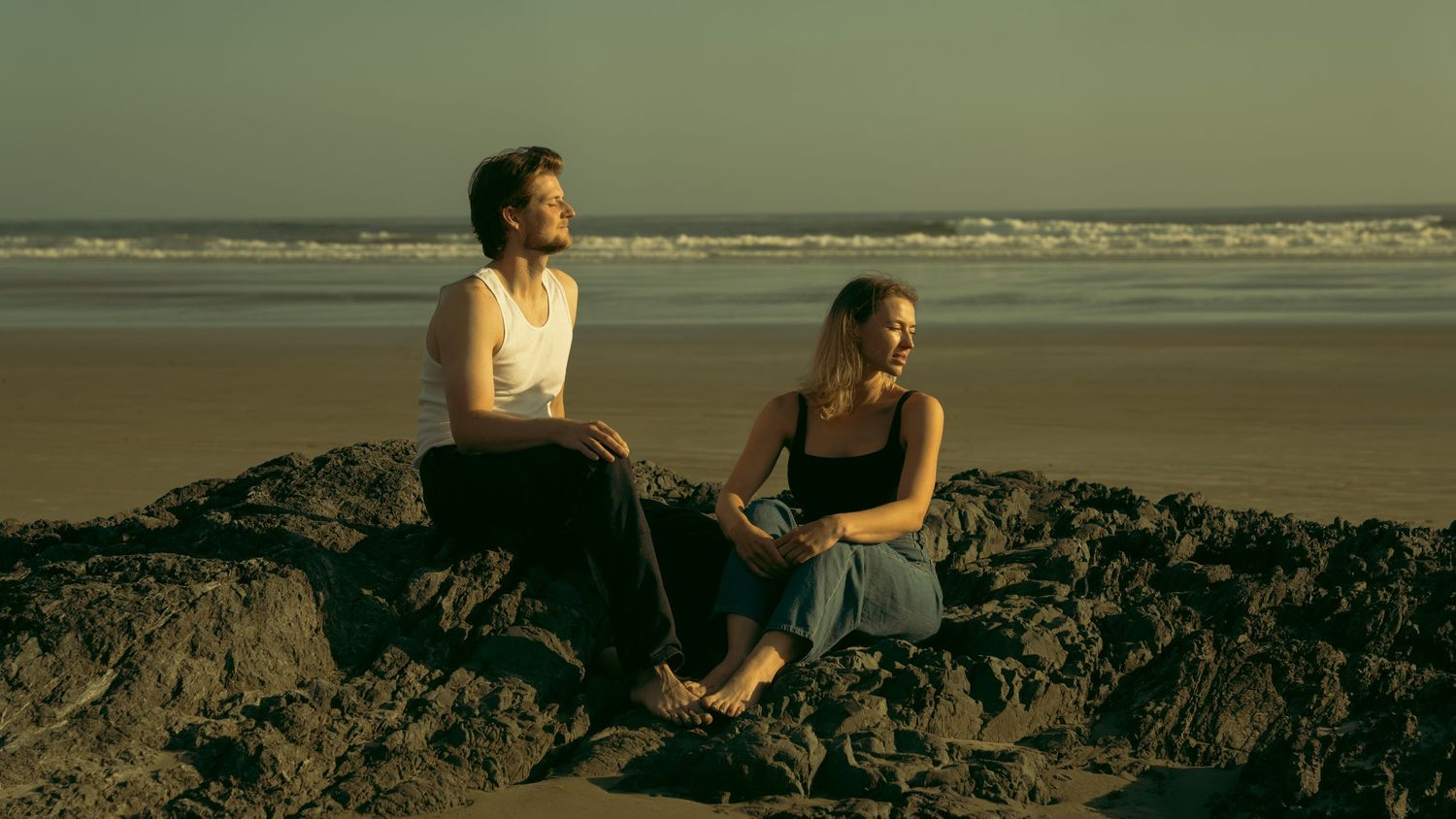 Two people sitting together on coastal rocks at dusk overlooking the ocean.