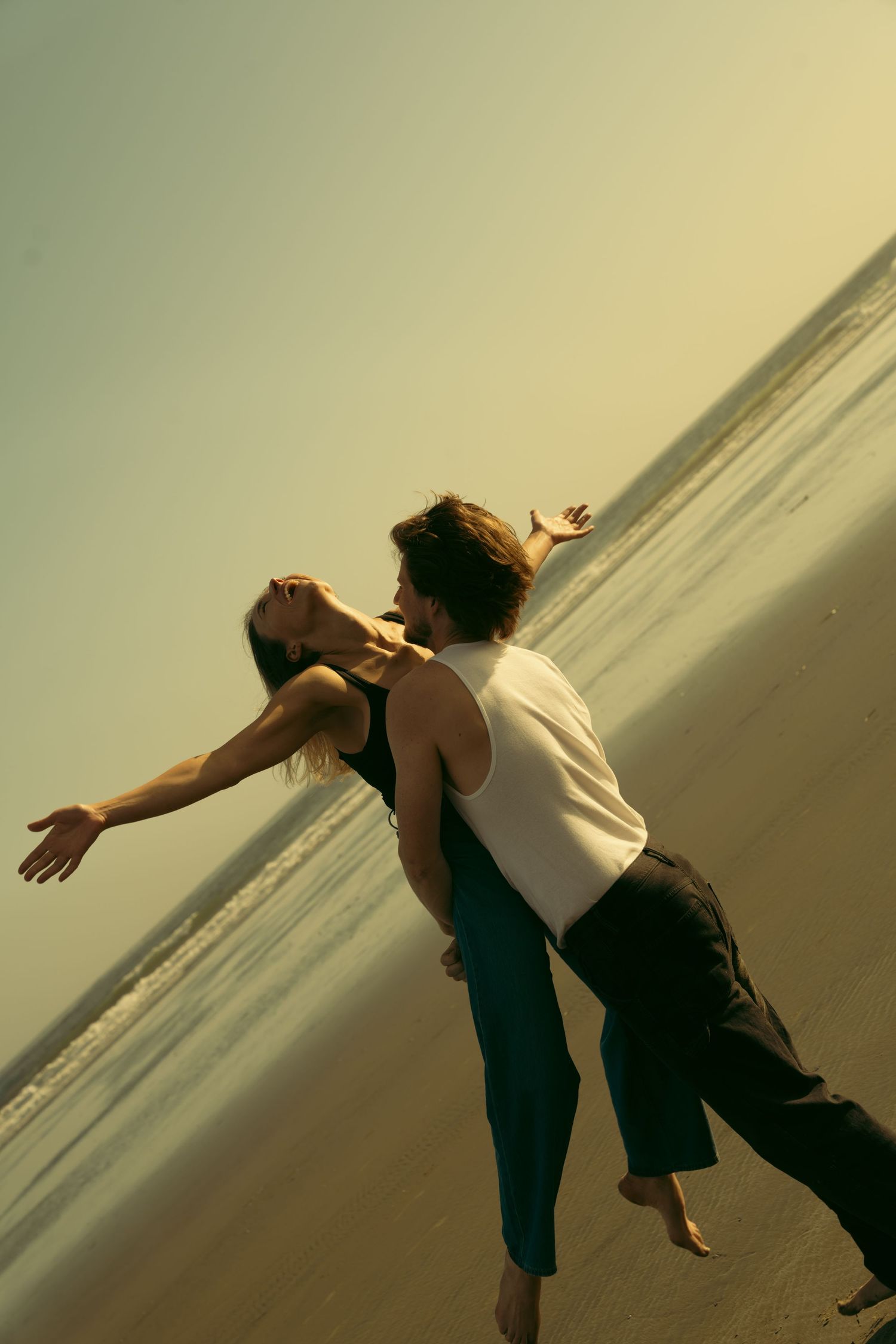 A couple dances playfully on a beach at sunset.