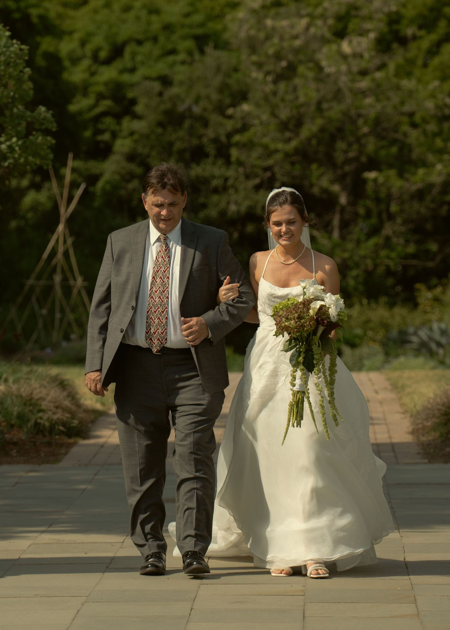 Bride in white gown holding bouquet walks down aisle with father in gray suit.