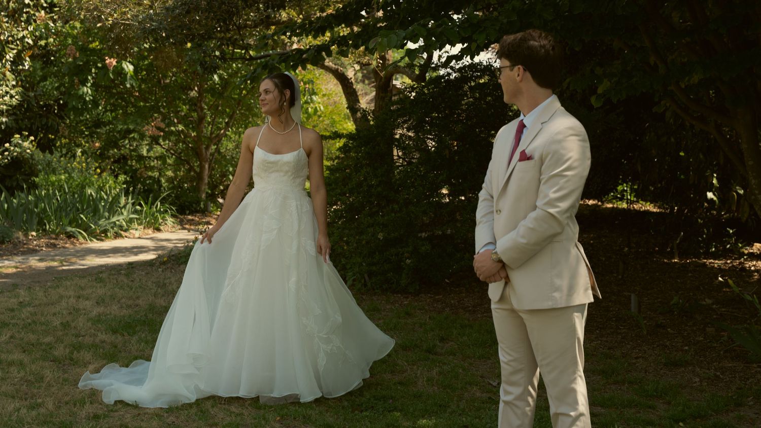 Bride in white gown and groom in tan suit share a romantic first look outdoors in a lush garden setting.