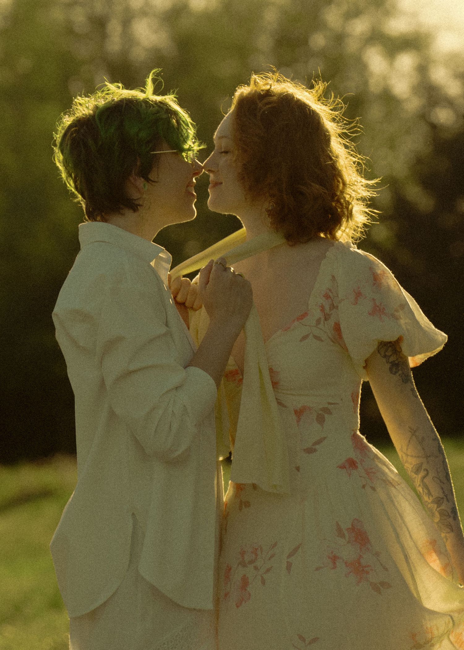 Two women share a romantic moment, nearly kissing in warm golden sunset light outdoors.