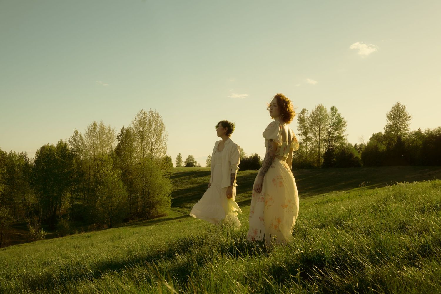 Two women in elegant white dresses walk through a lush green meadow at golden hour sunset.