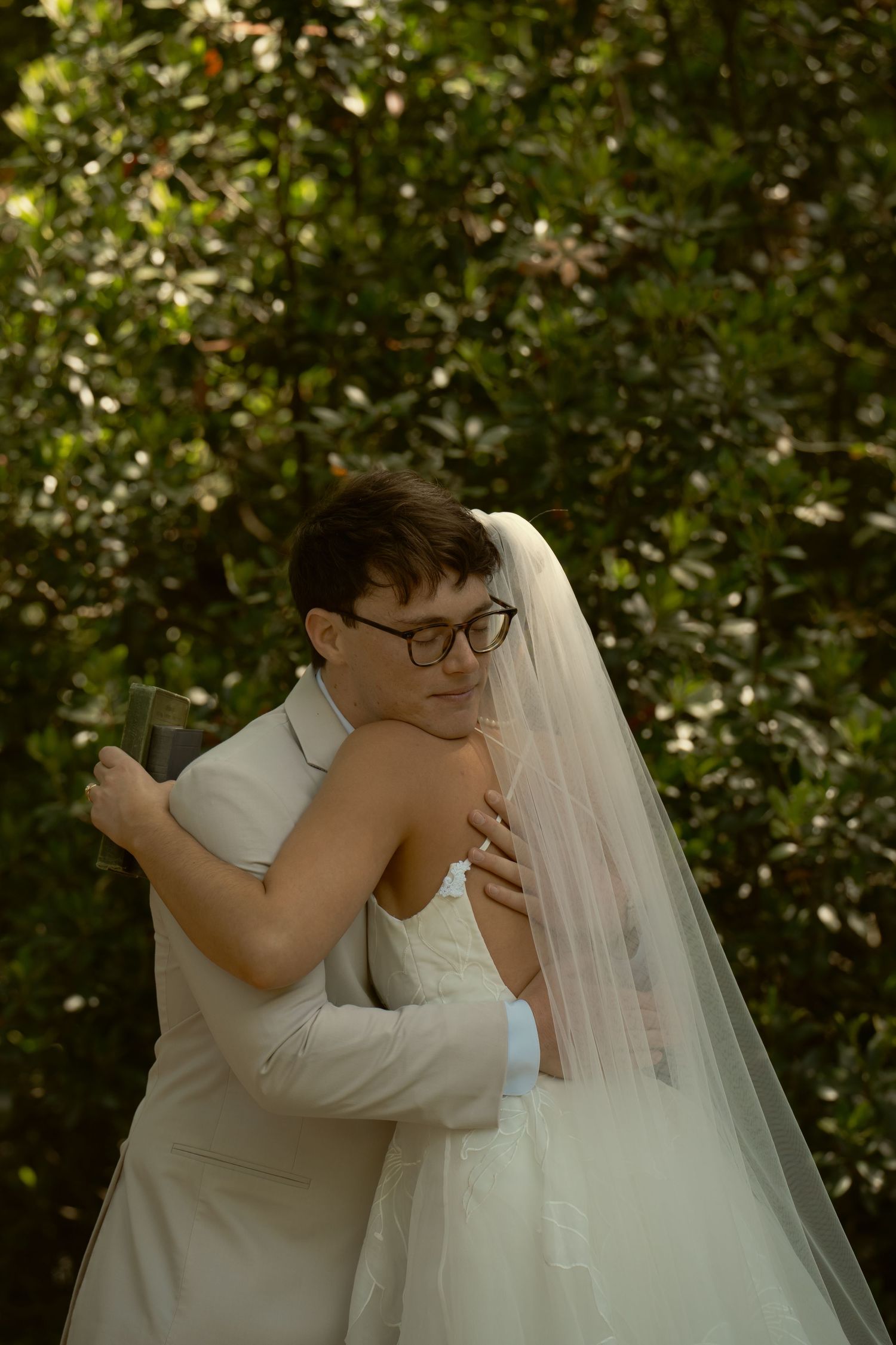 Groom in white suit embracing bride in veil tenderly among green garden foliage.