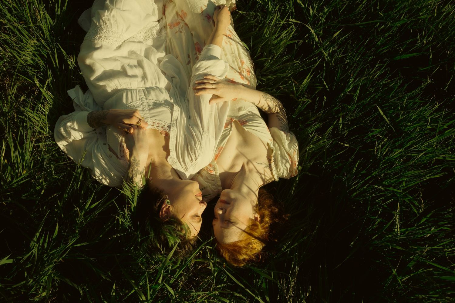 Two women in white dresses lying head-to-head on lush green grass in soft natural light.