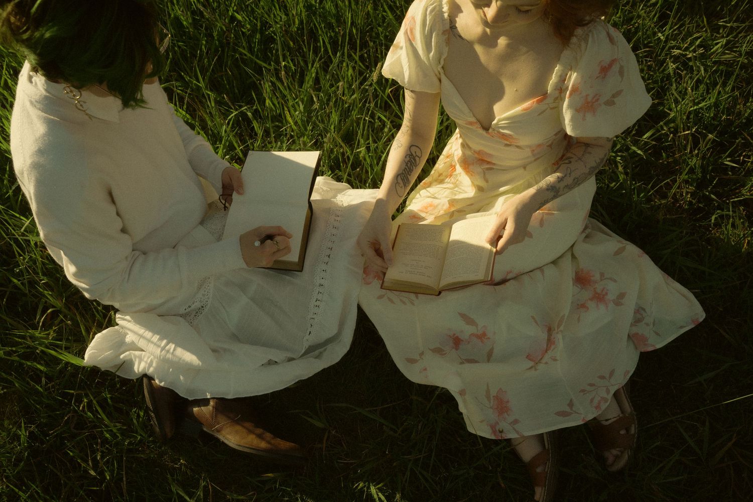 Two women in white floral dresses sit on green grass, reading books together in soft natural light.