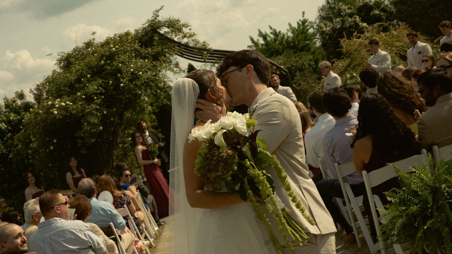 Bride and groom share romantic kiss at outdoor garden wedding ceremony with seated guests.