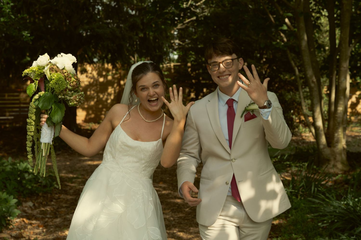 Newlyweds showing wedding rings, bride in white gown holding bouquet, groom in tan suit with pink tie, outdoors.