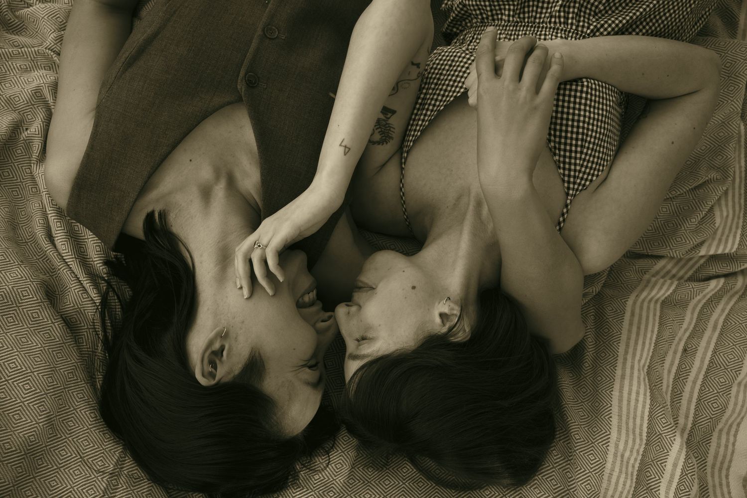 Two people lying close together on a bed in a sepia-toned intimate portrait photograph.