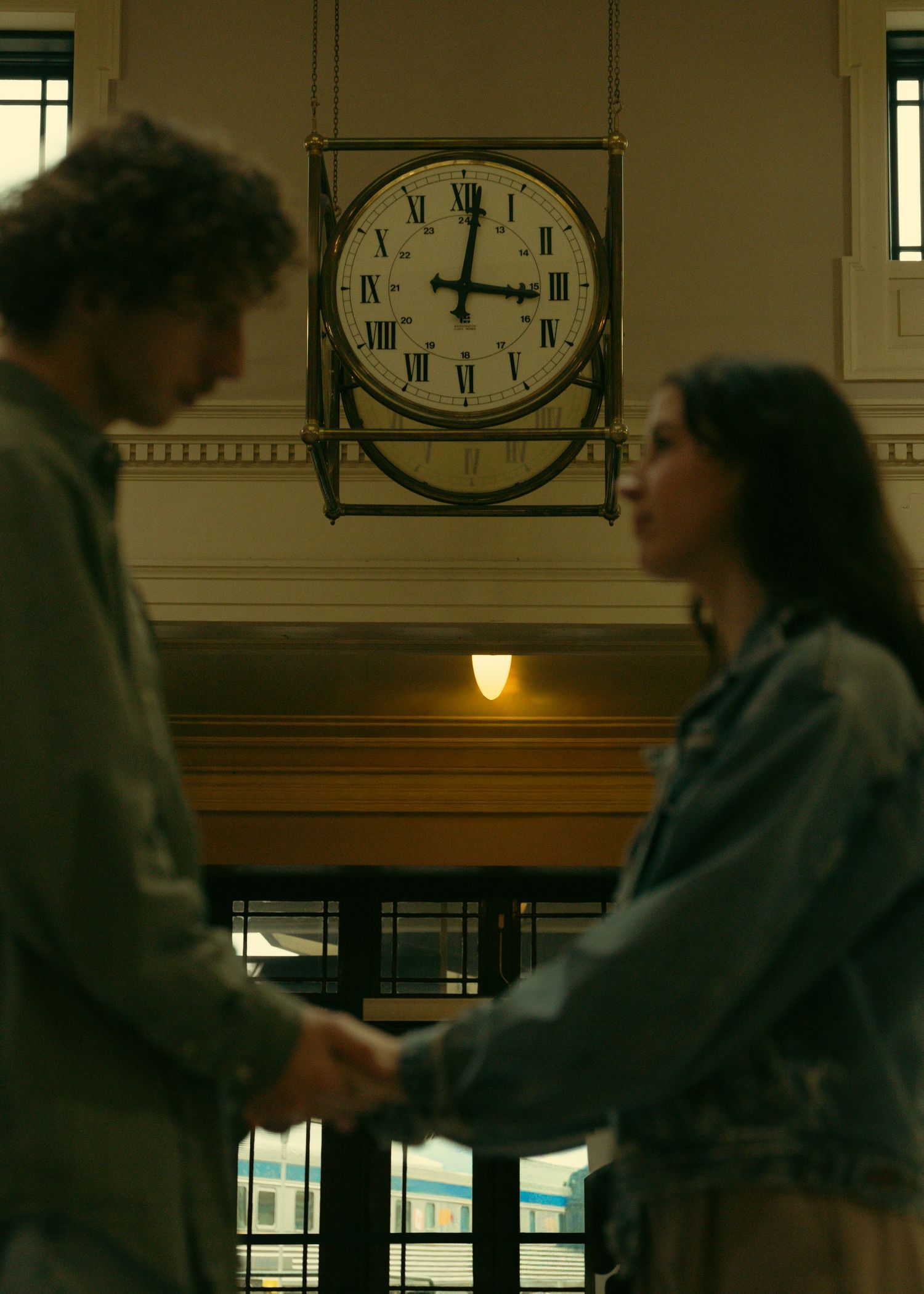 Two figures stand beneath a large clock in an ornate building interior.