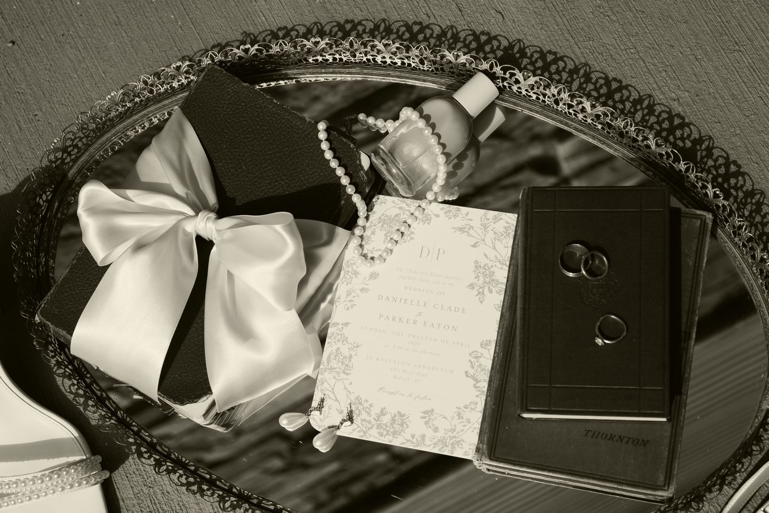 Wedding flat lay featuring rings, pearl necklace, invitation, and white bow on decorative silver tray.