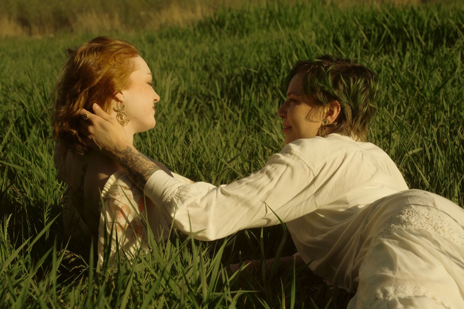 Two women share an intimate moment lying in tall green grass in warm golden sunlight.