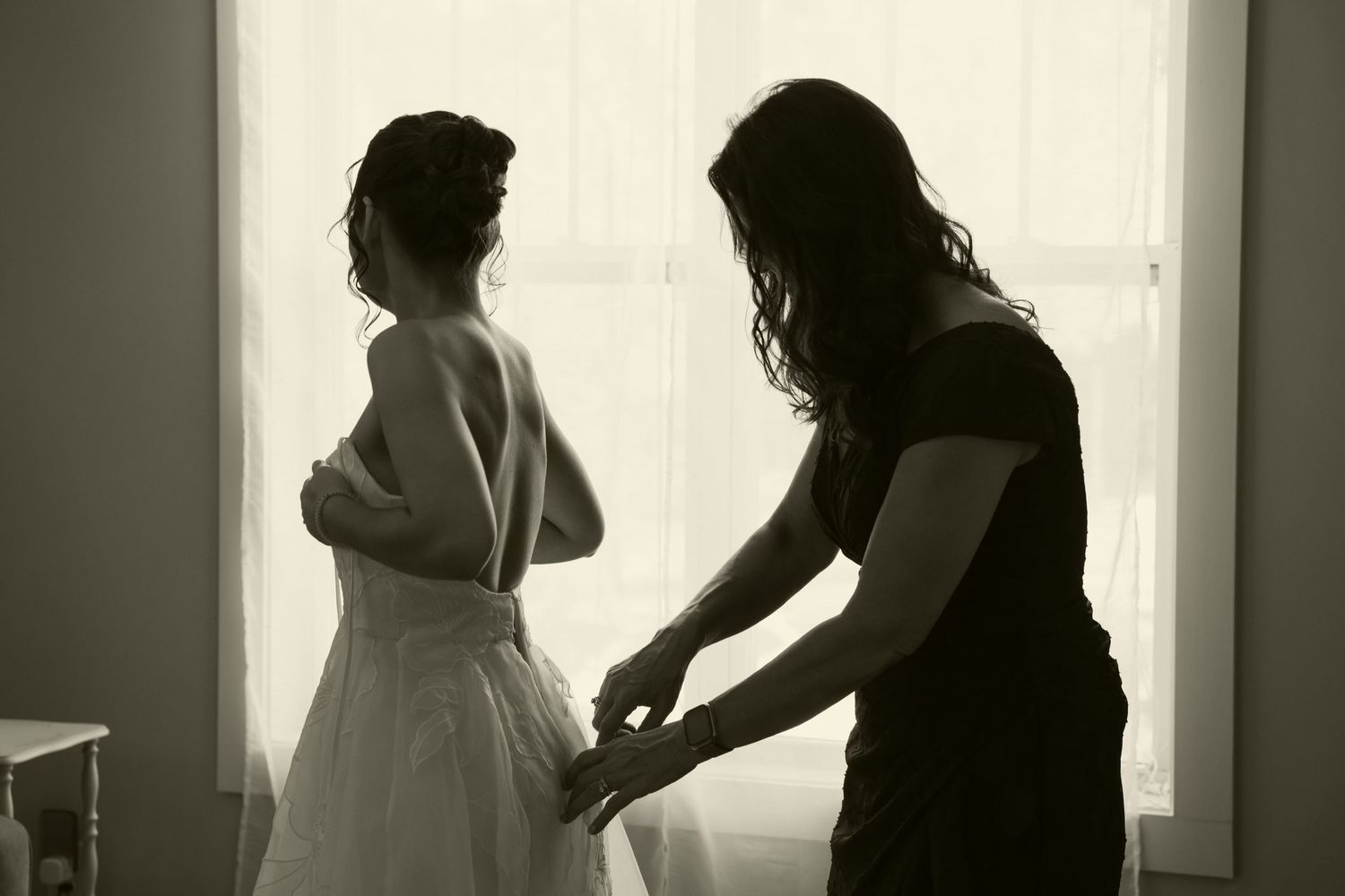 Black and white silhouette of bride being helped into wedding dress by assistant at window.