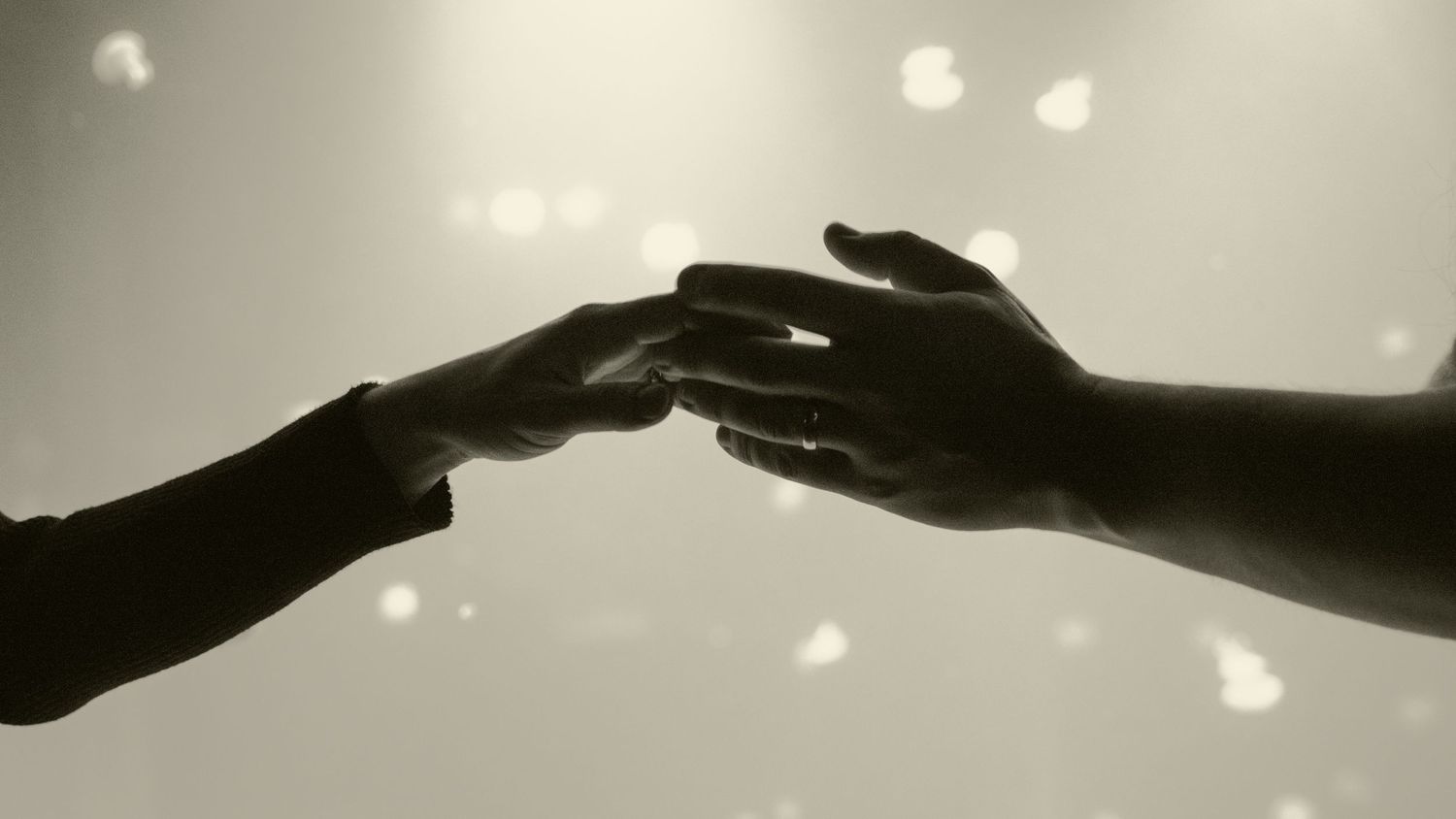 Two hands reaching toward each other in a tender touch, captured in soft black and white bokeh light.