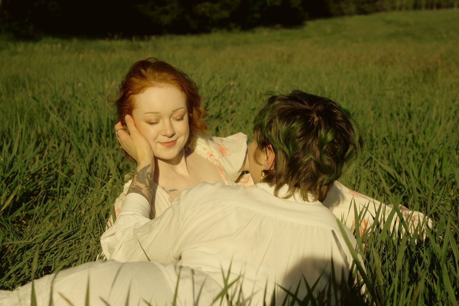 Two women lying in lush green grass sharing a tender romantic moment in warm natural sunlight.