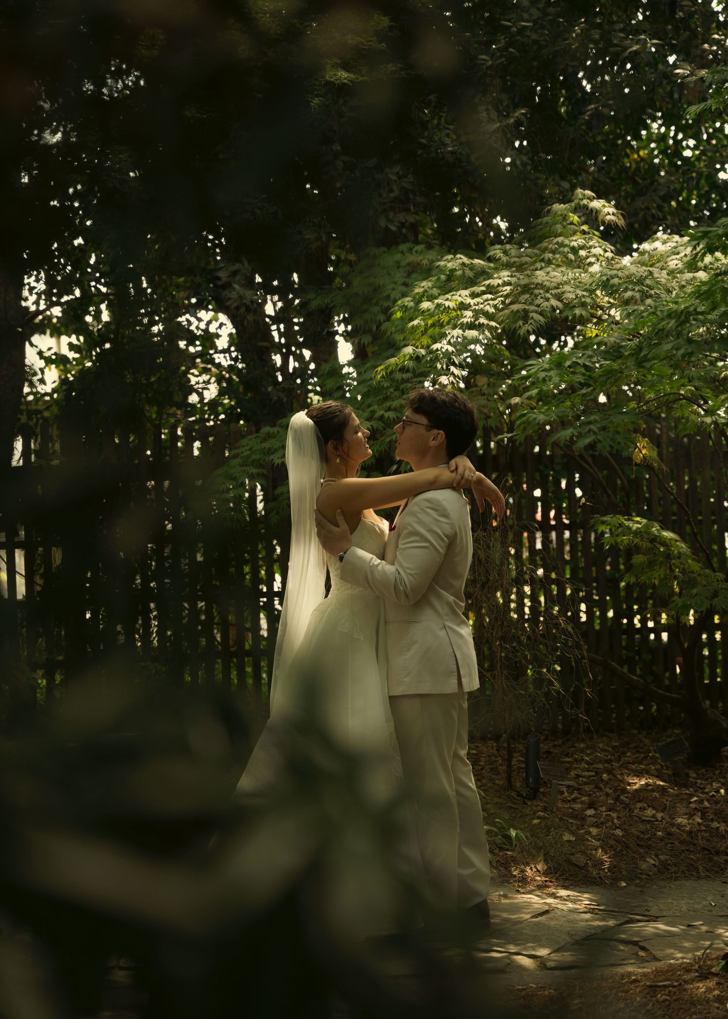 A bride and groom share a romantic embrace in a lush garden setting, surrounded by green trees and a wooden fence.