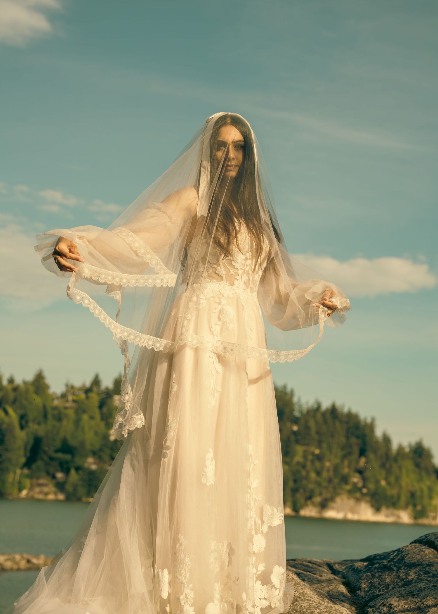 A figure in a flowing white lace dress and veil poses dramatically against a lakeside landscape at sunset.