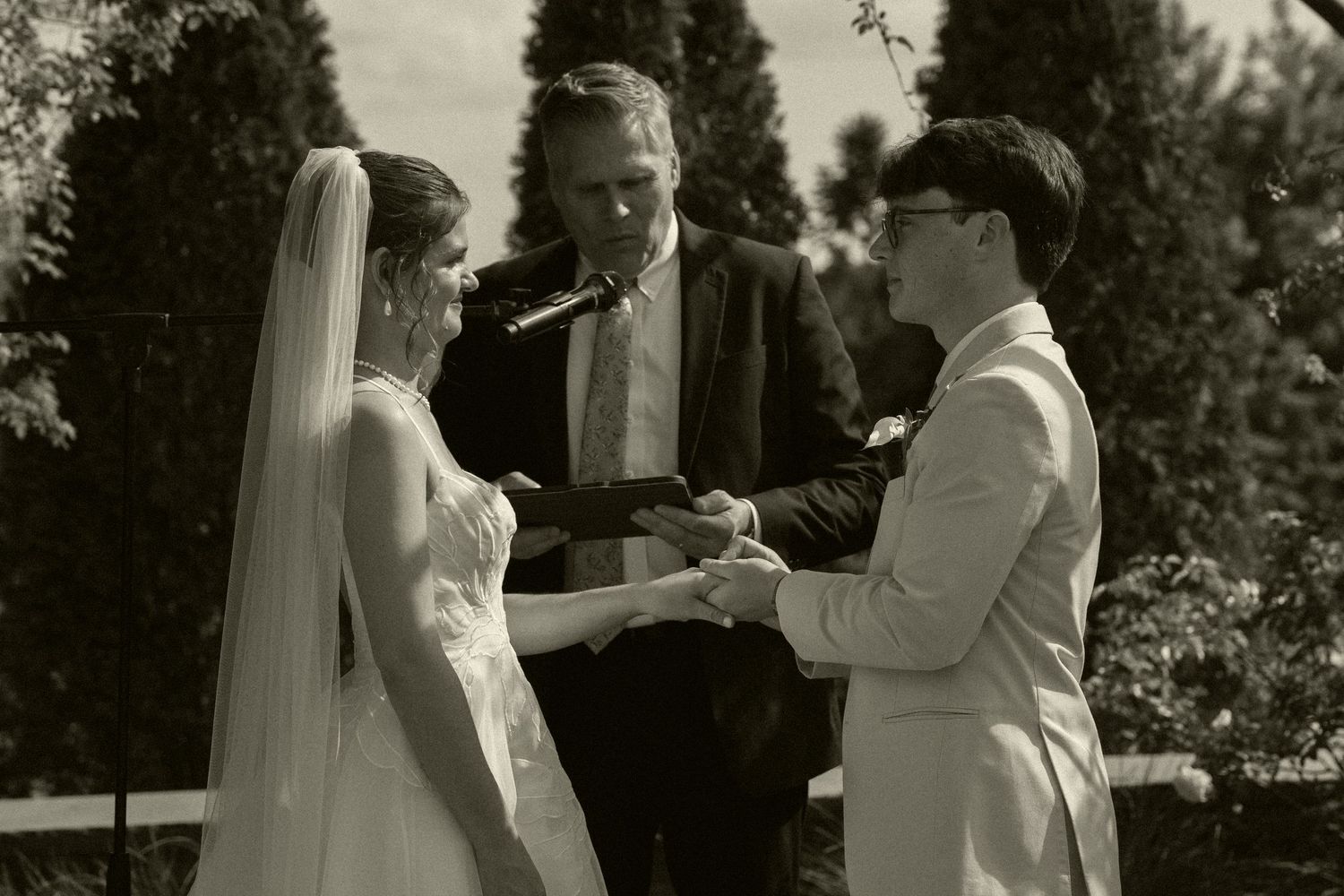 Black and white photo of bride and groom exchanging vows with officiant outdoors.